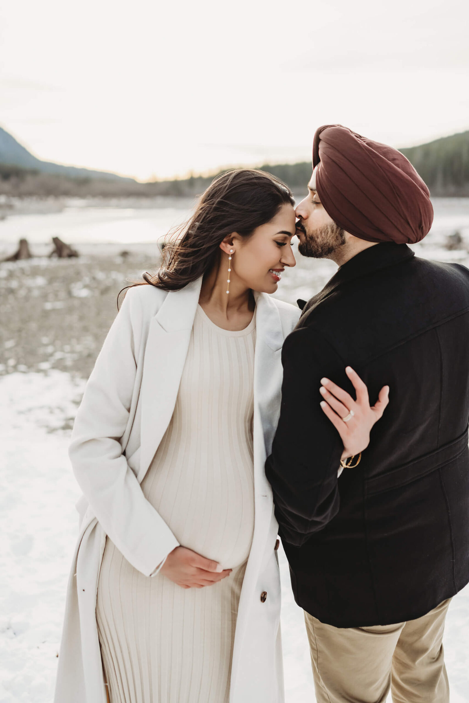 Expecting parents standing together in a snowy lake setting during a winter outdoor maternity session.