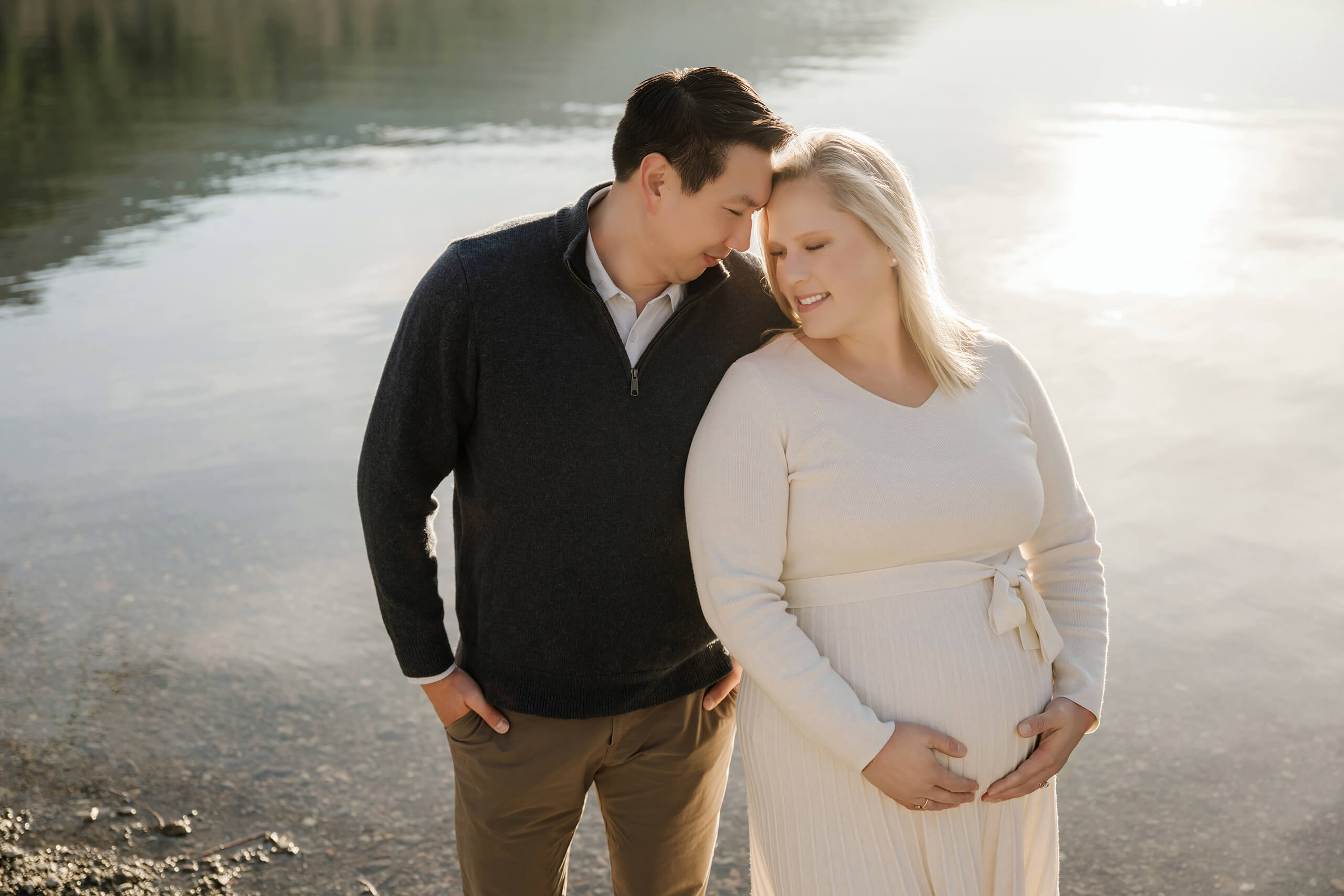 Expecting couple standing close together by the lake during a soft golden hour maternity session.