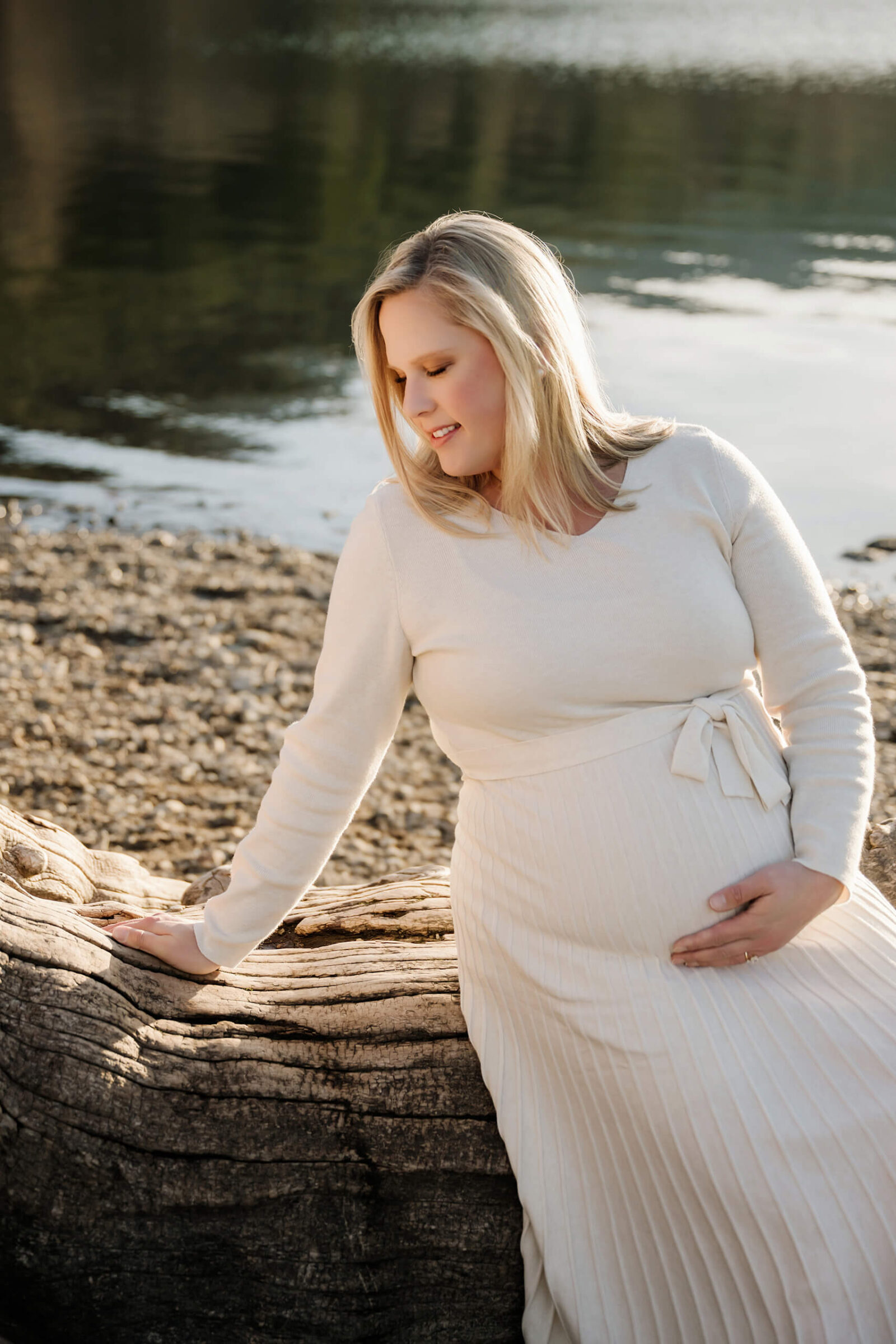 Expecting mother in a cream dress sitting by driftwood near the lake during an outdoor maternity session.