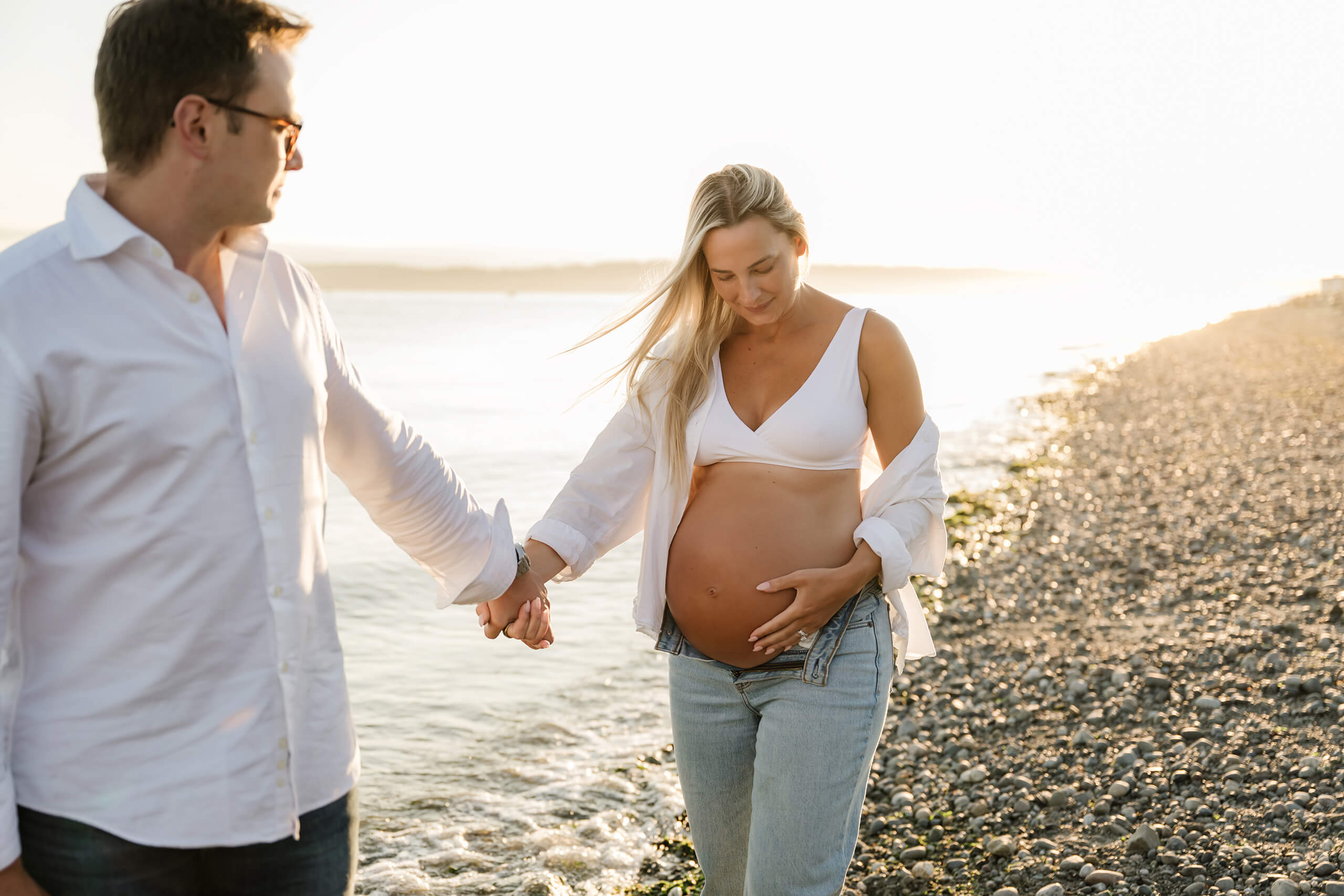 Expecting parents walking hand in hand by the water at Seattle Discovery Park during a golden-hour outdoor maternity session.
