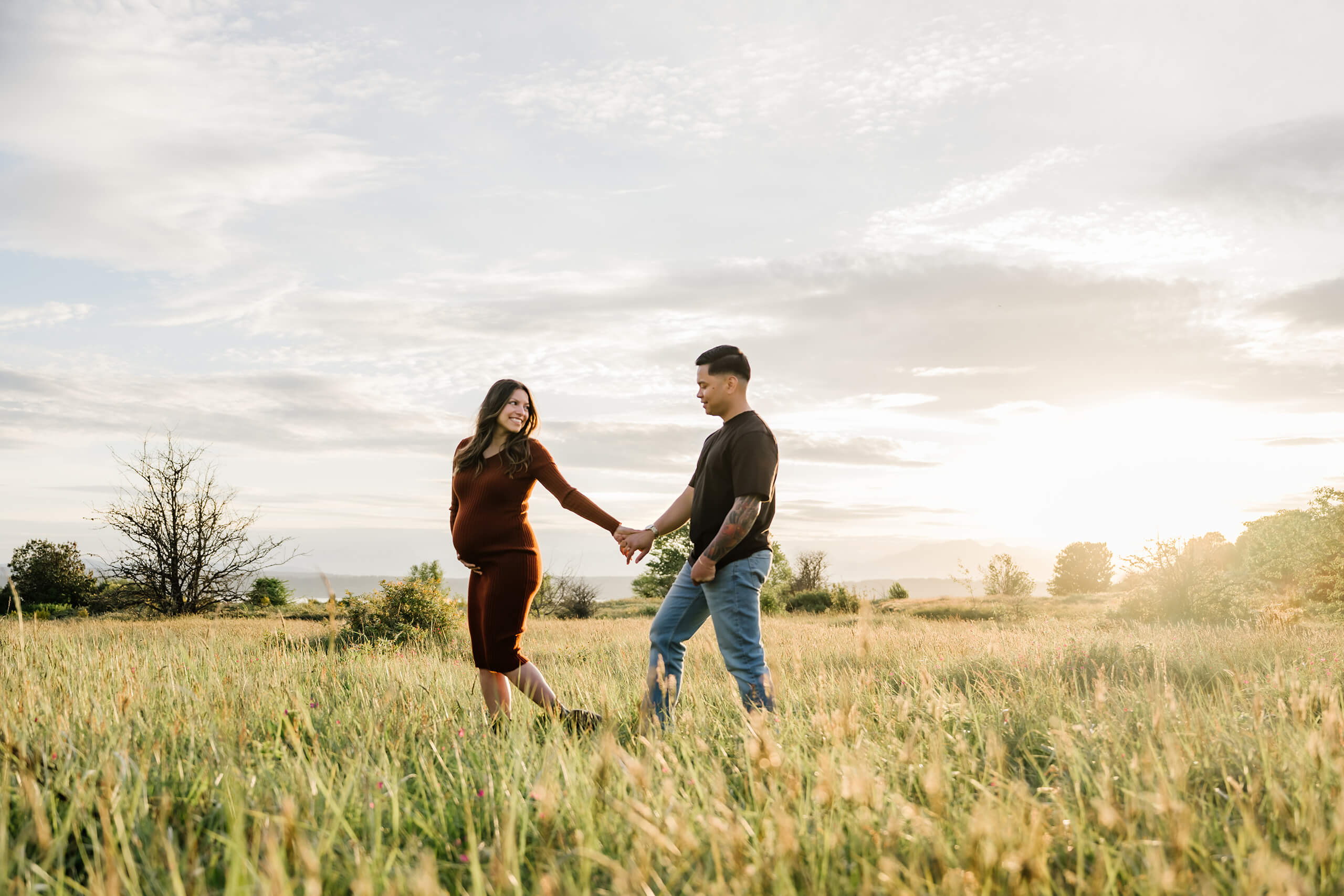 Expecting parents holding hands in a sunlit field during a natural outdoor maternity session at Seattle Discovery Park.