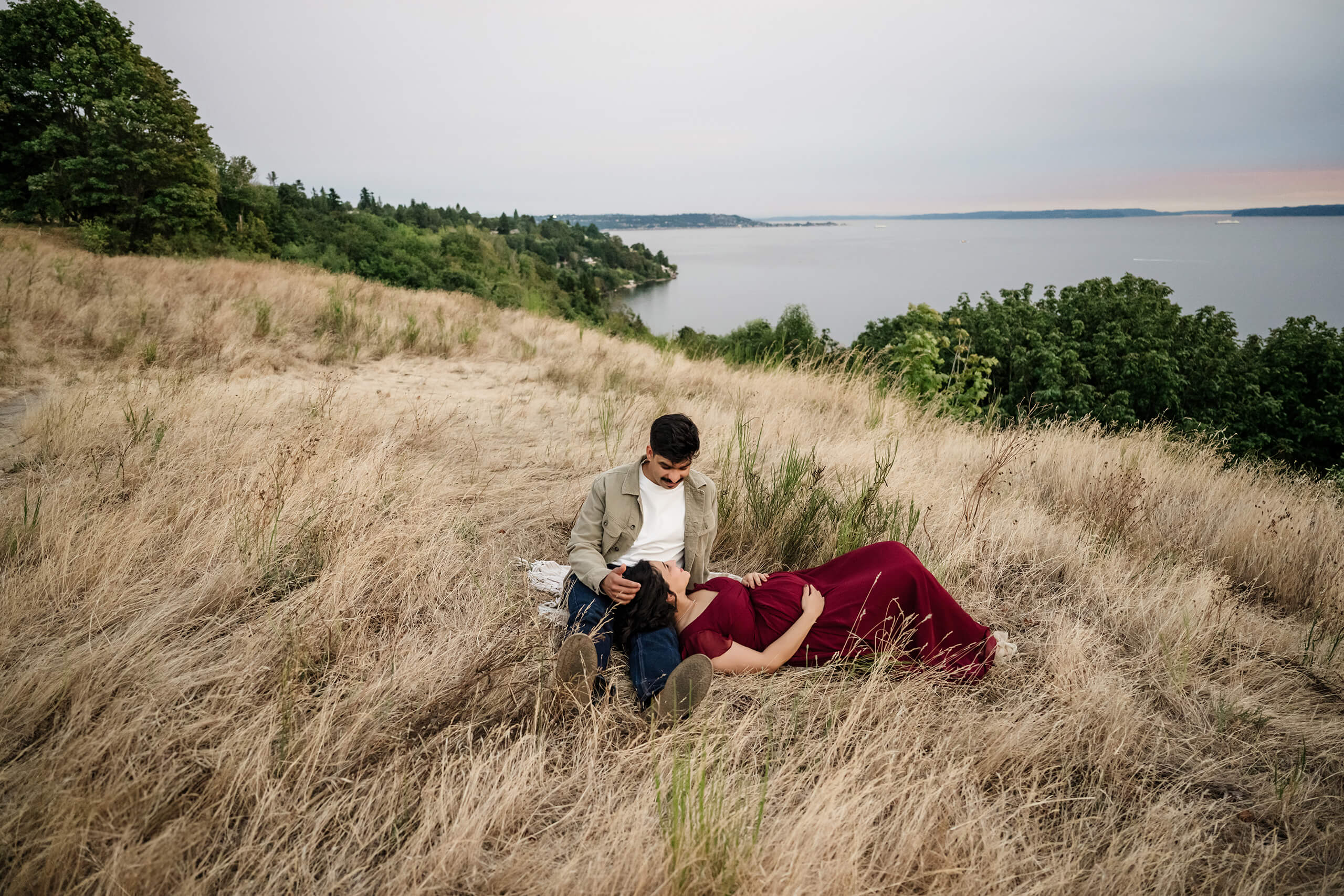 Expecting parents cuddling in a grassy hillside field with water views during an outdoor maternity session.