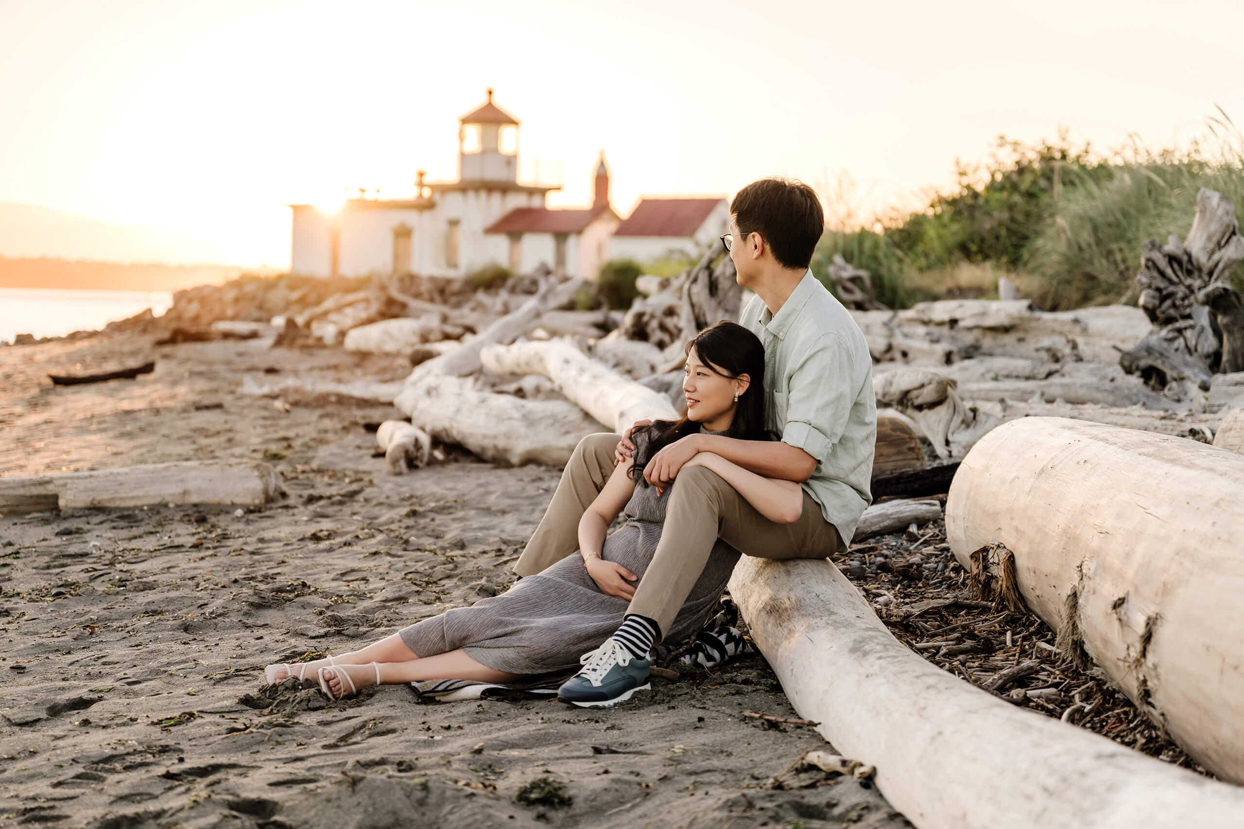 Expecting parents sitting together on a driftwood beach near a lighthouse during a golden-hour outdoor maternity session in Seattle.