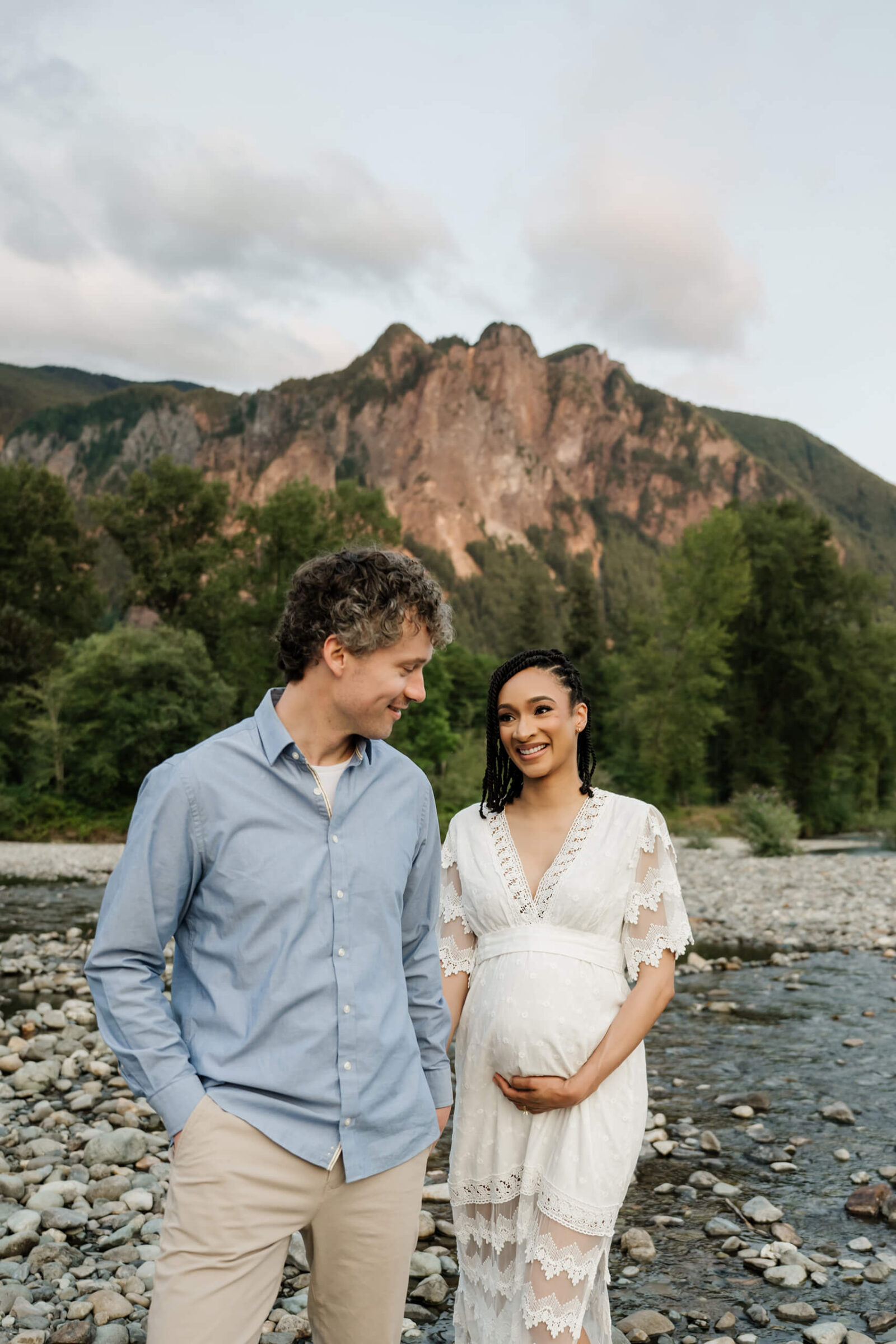 Expecting parents standing together by a rocky river with mountain views during a natural outdoor maternity session near Seattle.