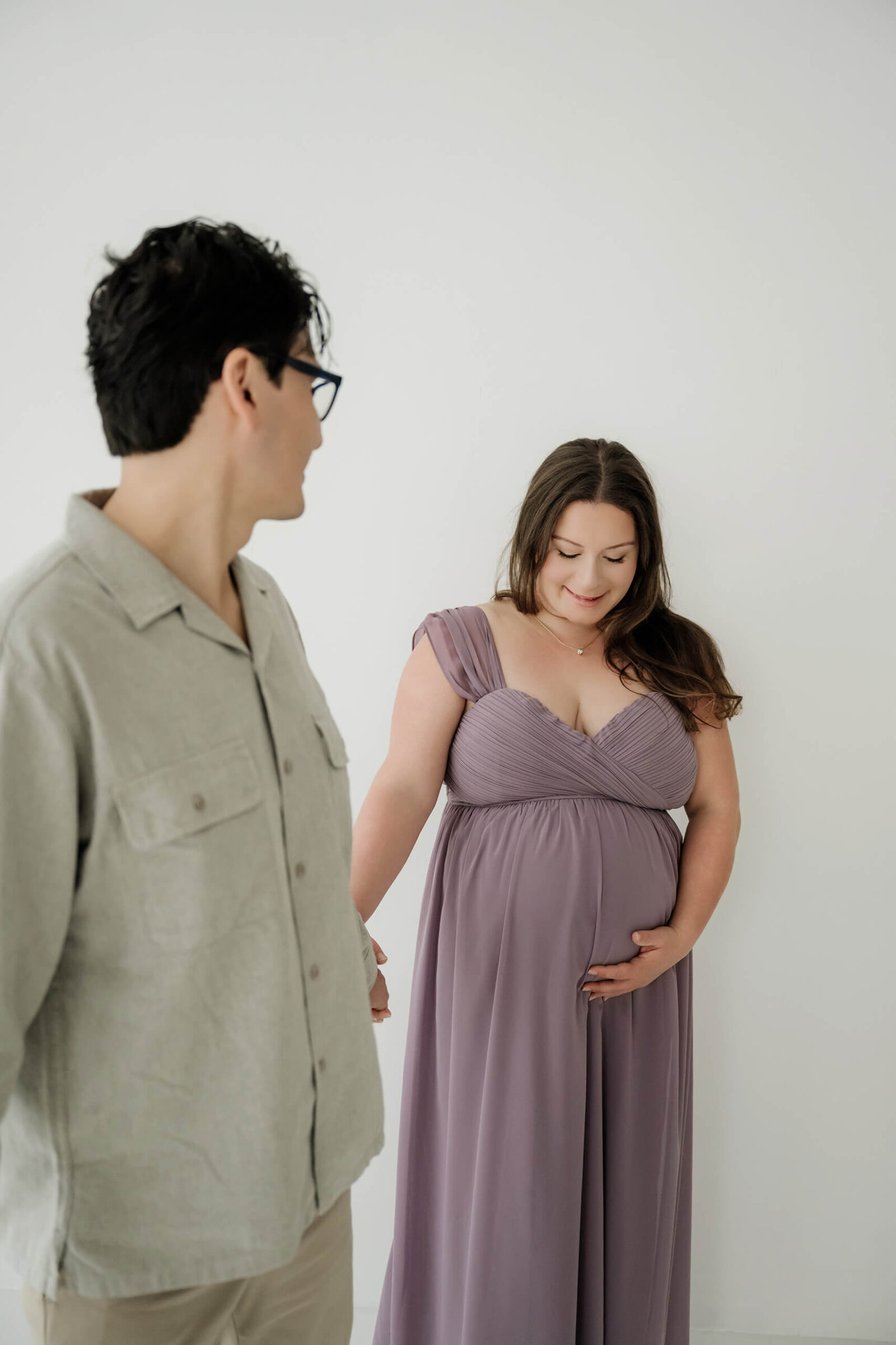 Expecting mom holding her belly while her partner looks on during a relaxed studio maternity session.