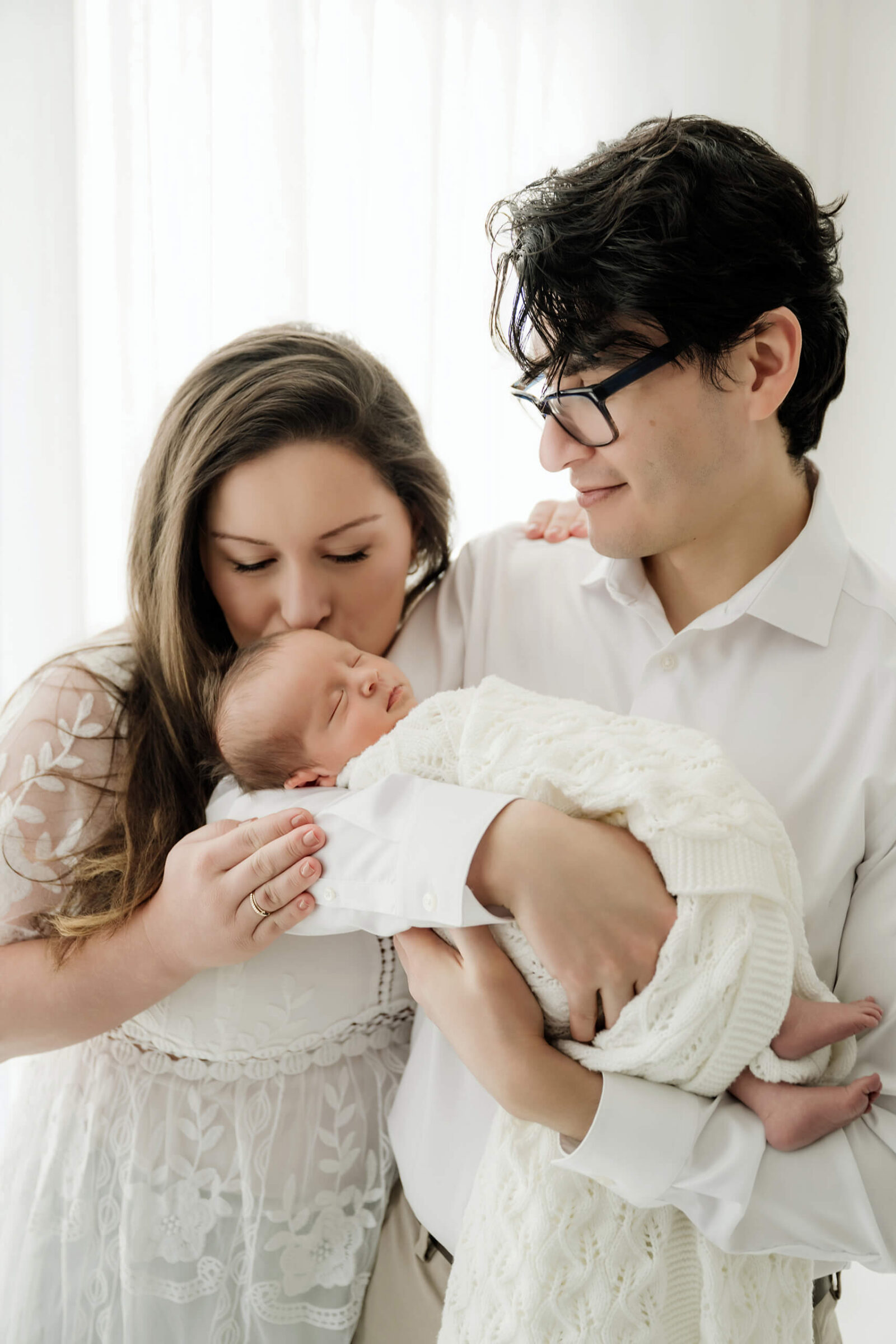 Parents holding their sleeping newborn baby in a cream knit blanket during a light-filled studio newborn session.