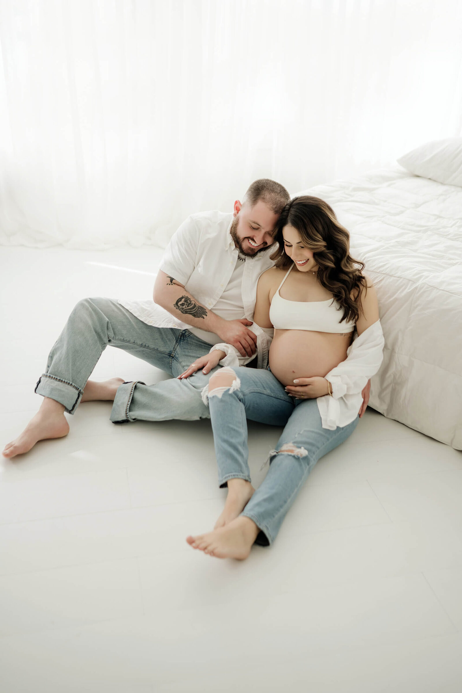 Expecting parents sitting together in a bright white studio during a relaxed maternity session.