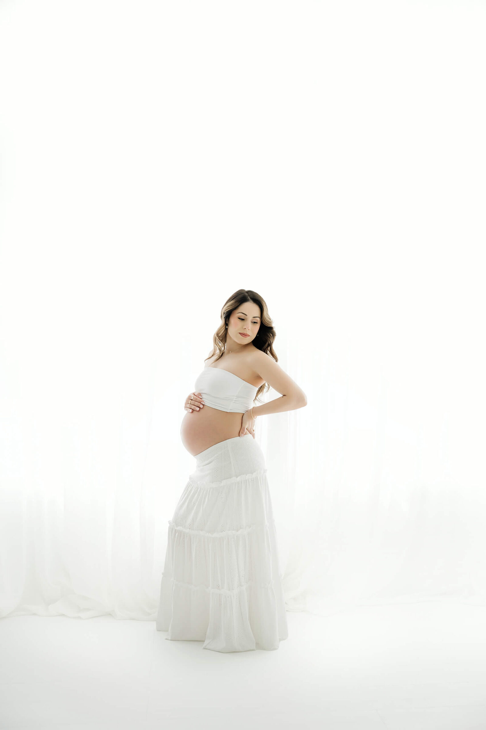 Expecting mom in a white skirt gently holding her baby bump during a timeless studio maternity session.