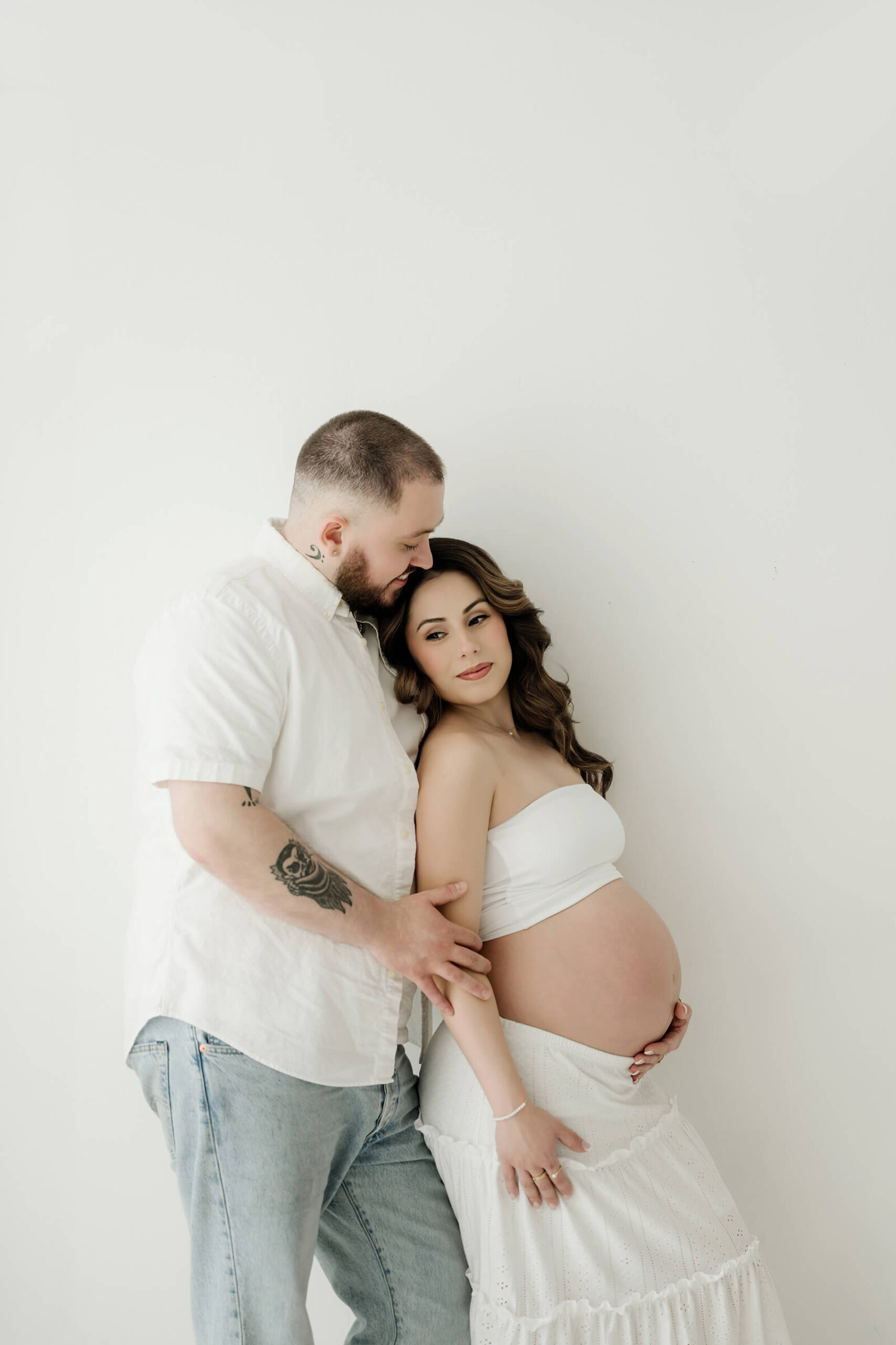 Expecting parents standing together in a clean white studio for a calm, natural maternity portrait.