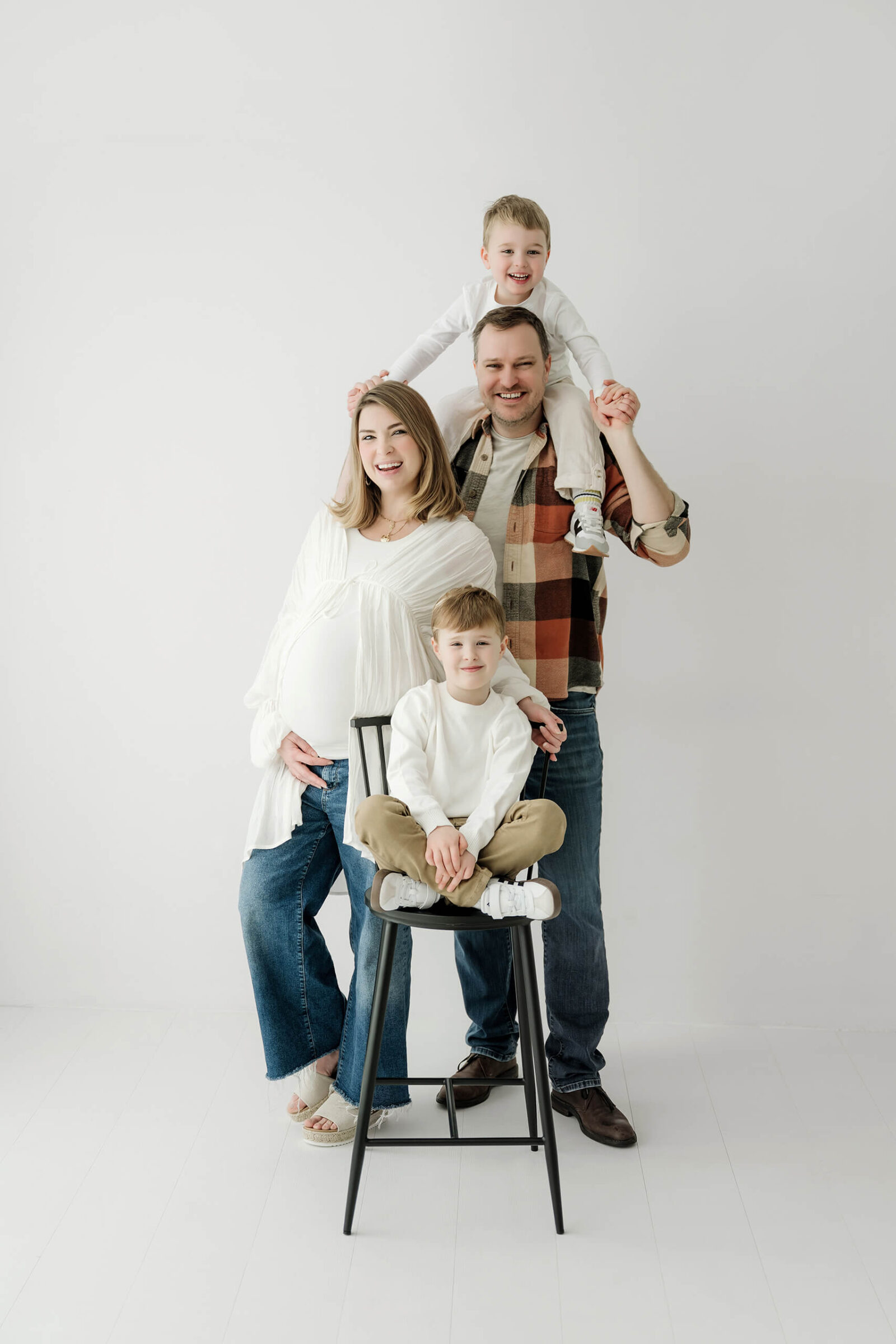 Growing family smiling together in a bright white studio during a natural maternity session with siblings.
