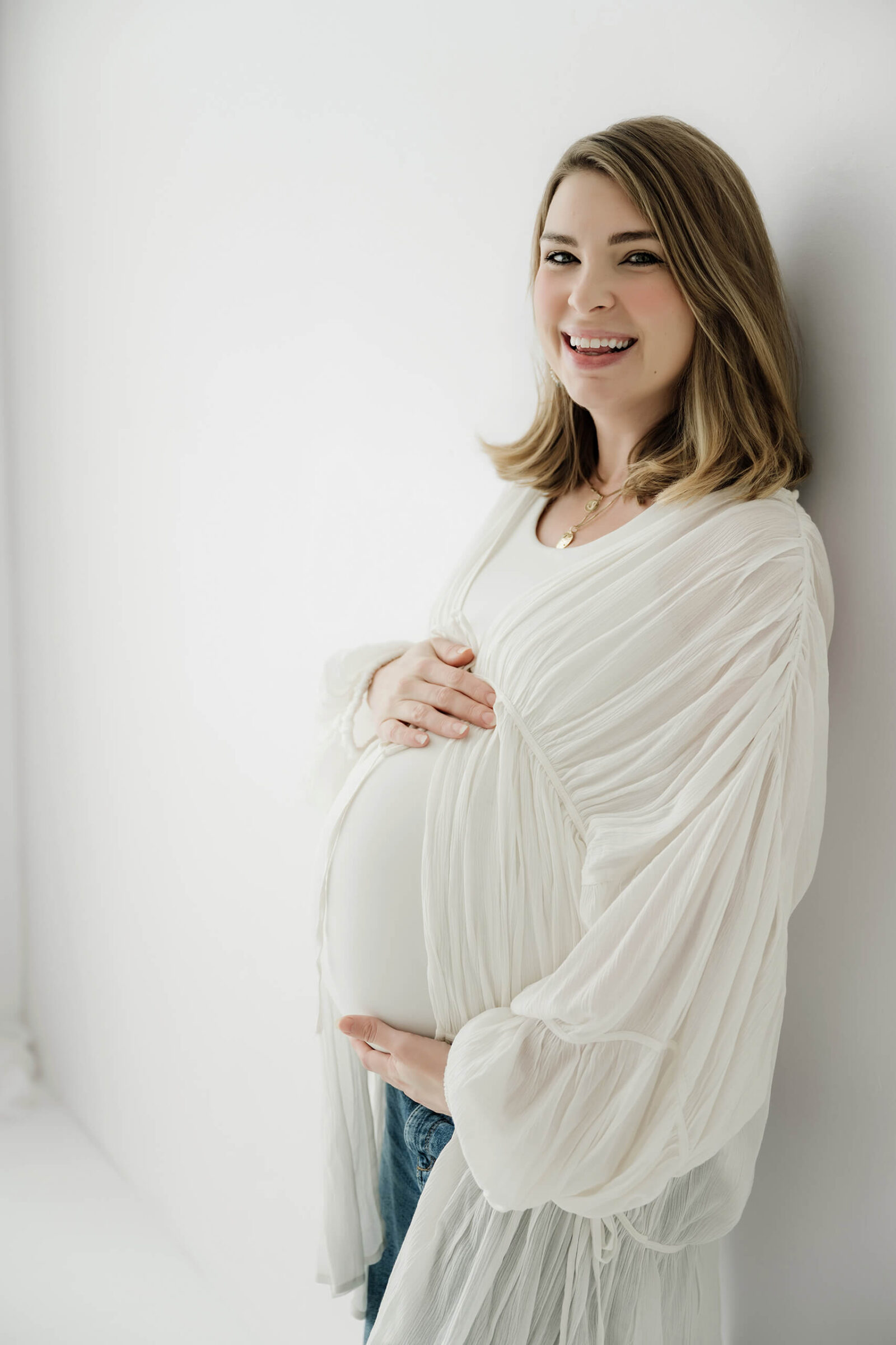 Expecting mom smiling and holding her baby bump during a soft white studio maternity session.