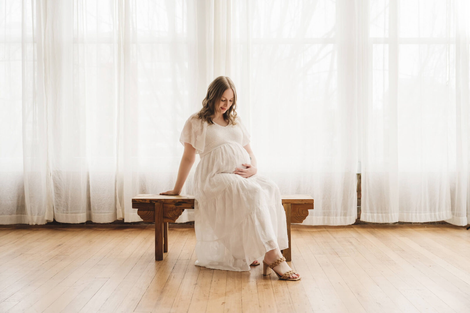 Expecting mother in a white dress sitting on a wooden bench during a Seattle studio maternity session.