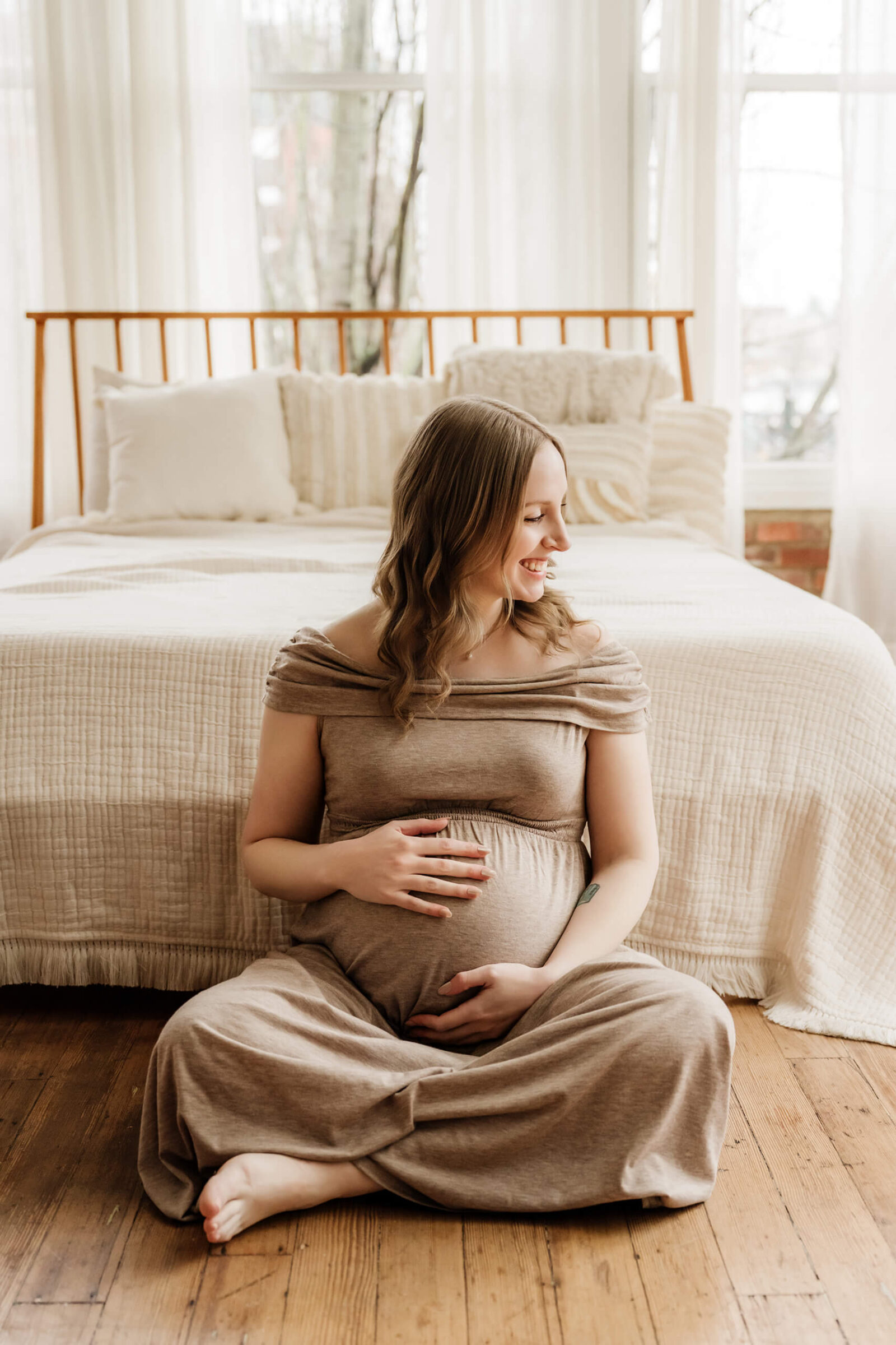 Expecting mother sitting on the studio floor in front of a neutral bed during a relaxed maternity session.