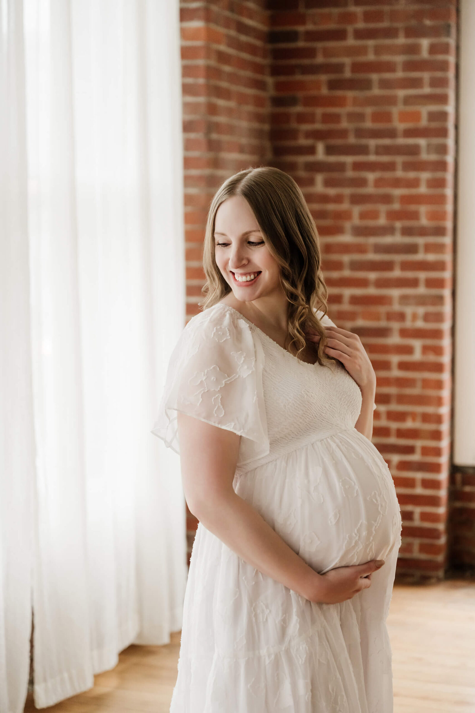 Expecting mother in a white dress smiling by soft curtains and brick walls during a studio maternity session.