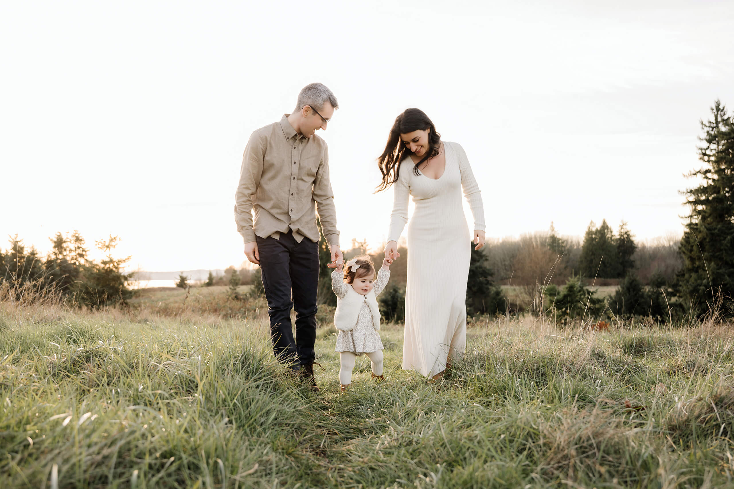 Parents walking hand in hand with their daughter through a grassy field at sunset.