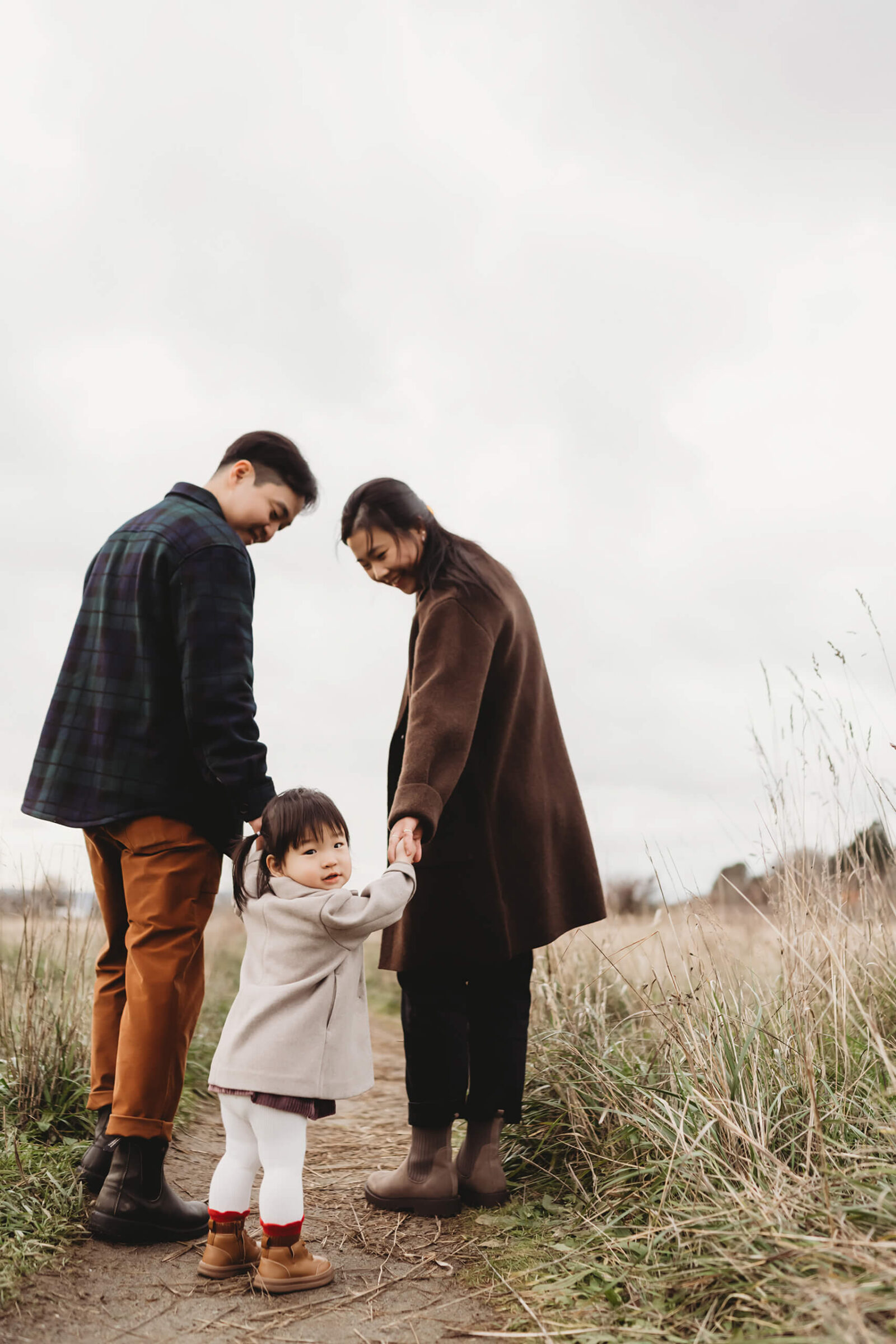 Parents holding their toddler’s hands while walking through tall grass during an outdoor family photo session.