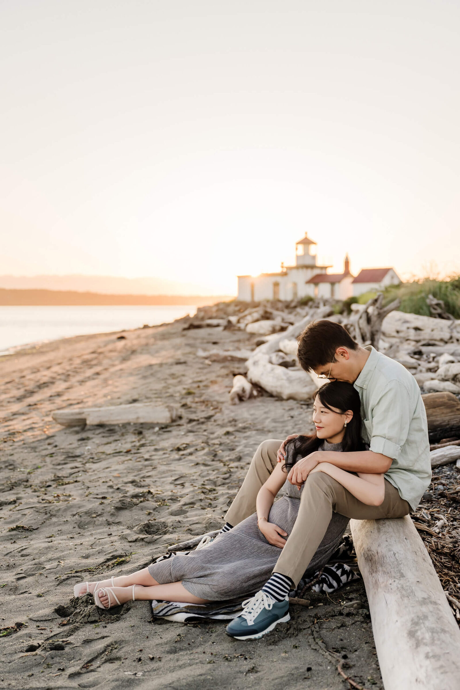 Pregnant mother leaning against her partner on a Seattle-area beach with a lighthouse in the background.