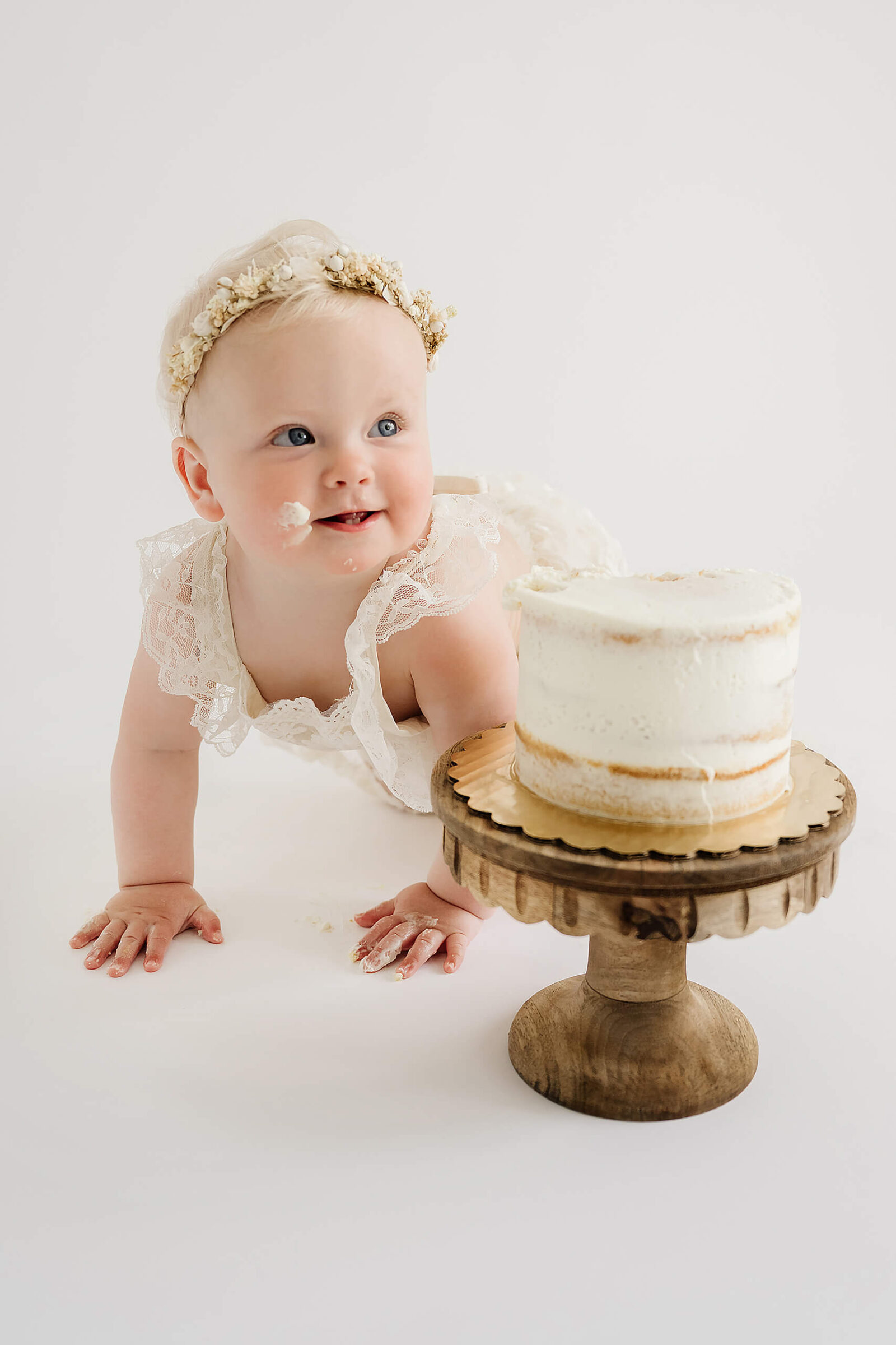 One-year-old crawling beside a white birthday cake with frosting on her face in a clean studio cake smash setup.