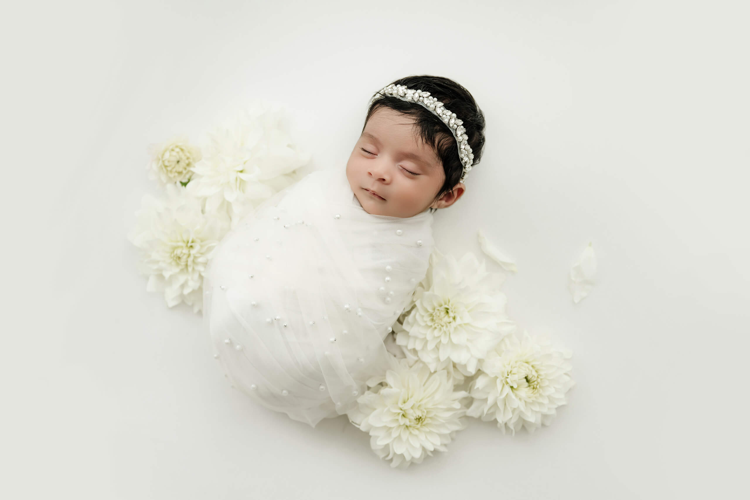 Sleeping newborn wrapped in white fabric surrounded by white flowers in a Bellevue newborn photography studio.