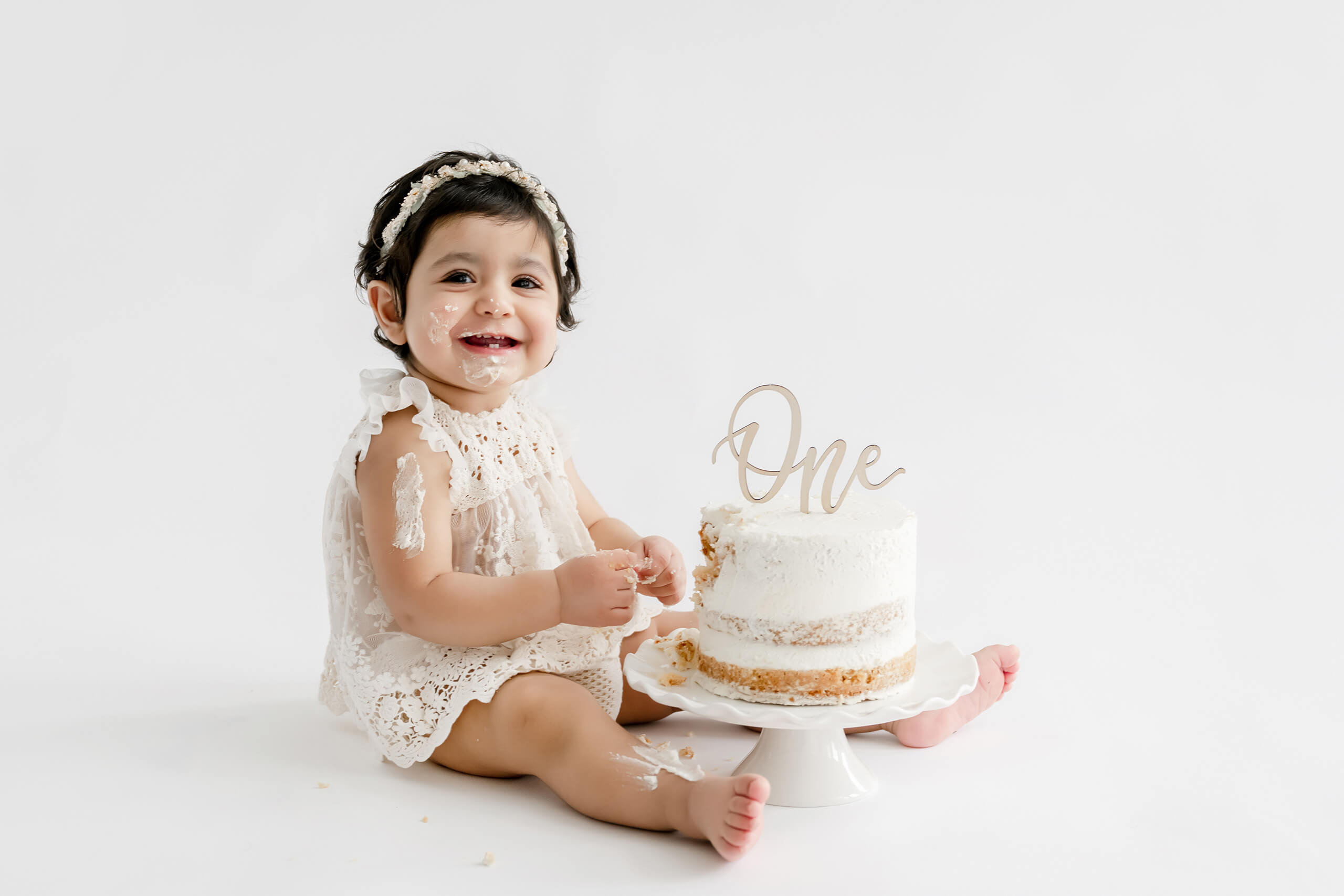 Smiling one-year-old sitting beside a white birthday cake during a clean studio milestone session.