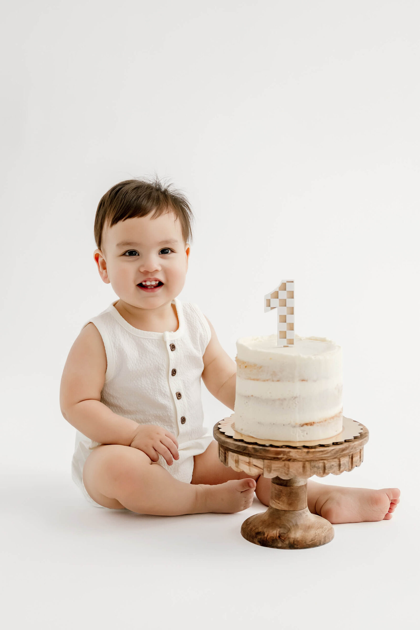Smiling one-year-old sitting beside a white birthday cake during a clean studio milestone session.