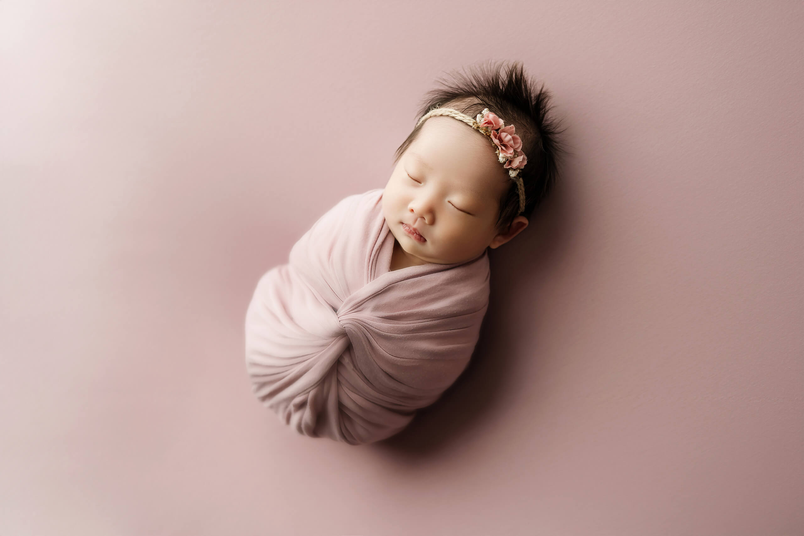 Sleeping newborn wrapped in pink fabric with a floral headband during a studio newborn session.