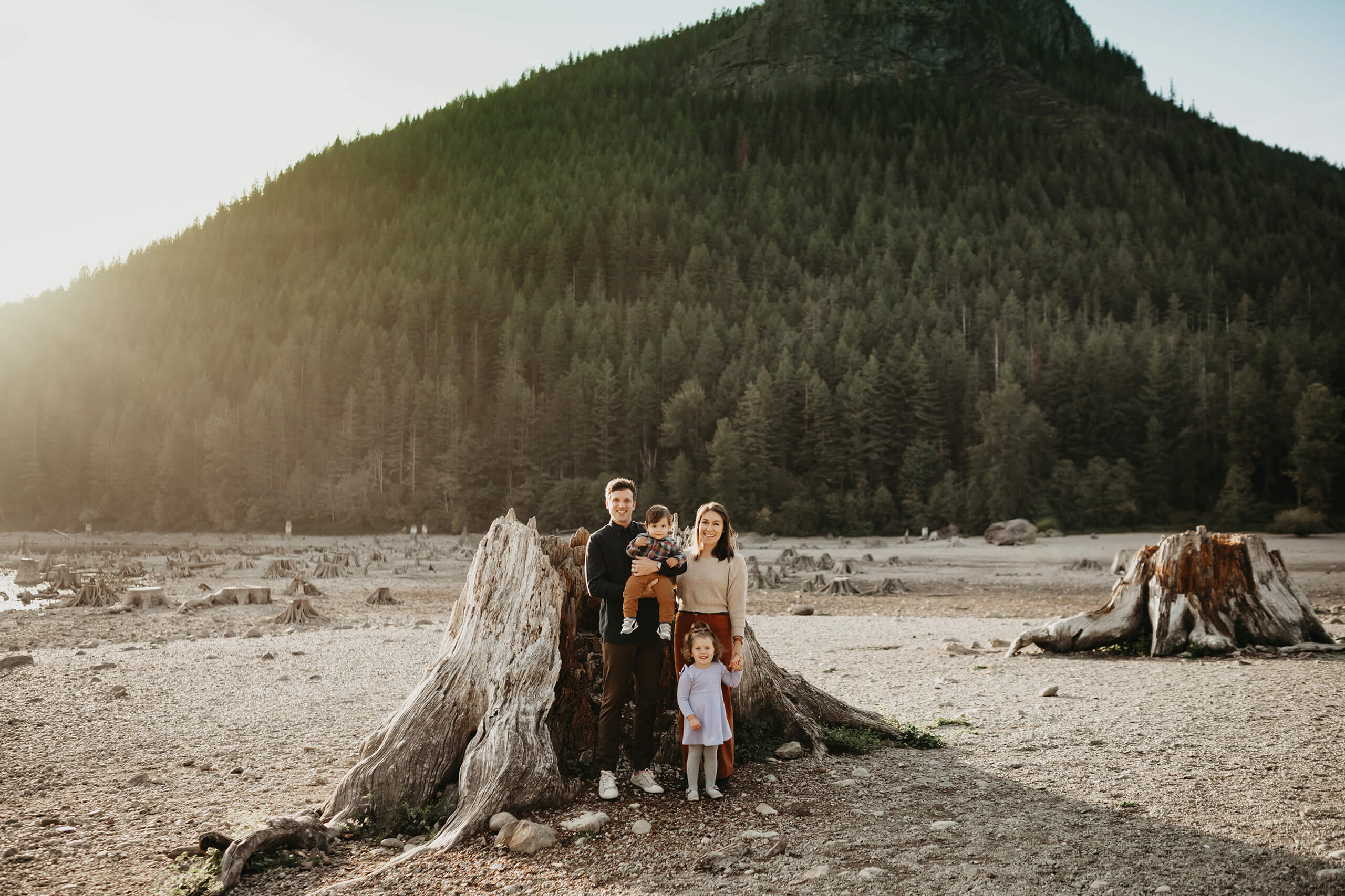 Family of four standing beside a large tree stump with forested mountains in the background.