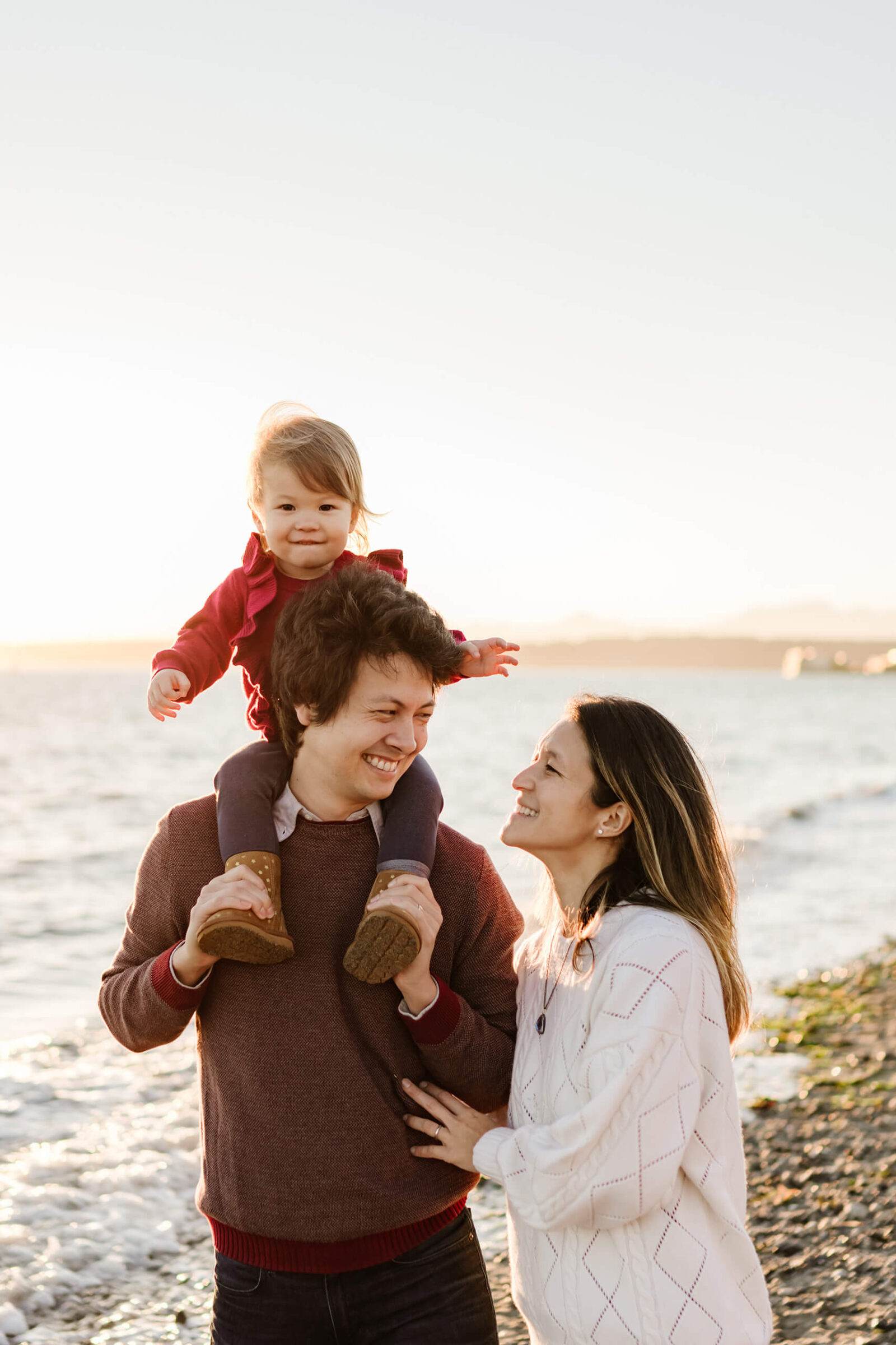 Toddler sitting on dad’s shoulders while mom smiles beside them during a sunset beach family photo session.