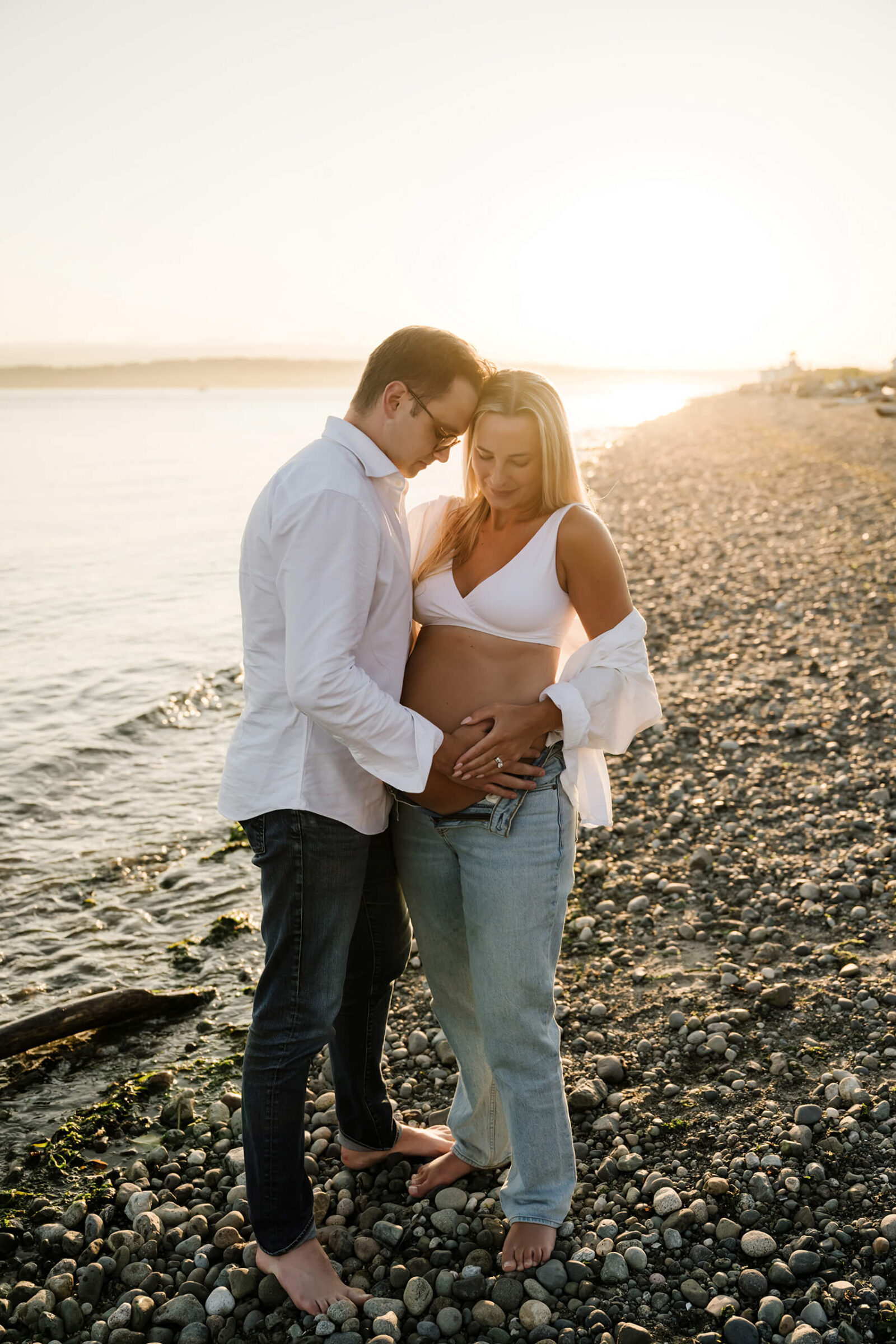 Expecting couple standing together on a rocky beach at sunset with their hands on the baby bump.