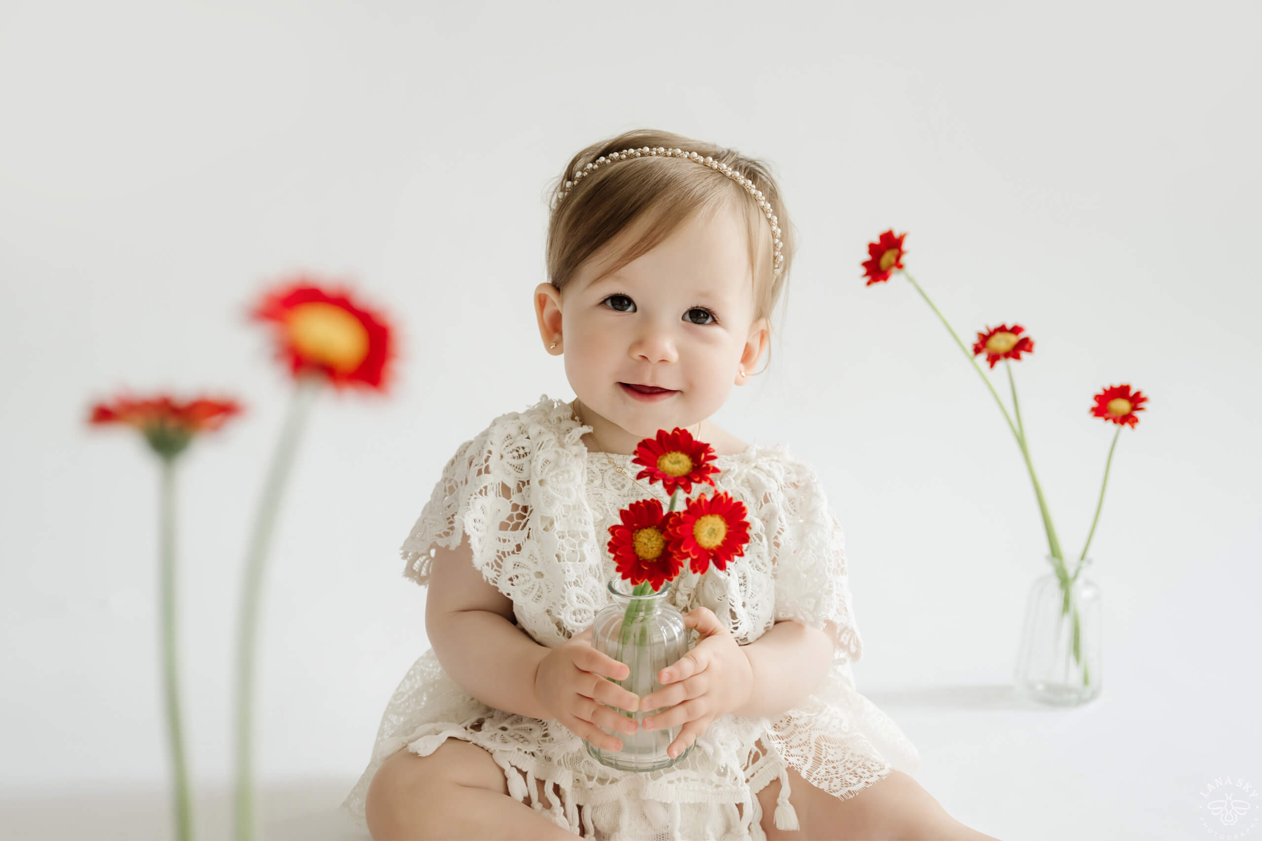 Baby in a white lace dress holding red flowers during a simple studio milestone portrait.
