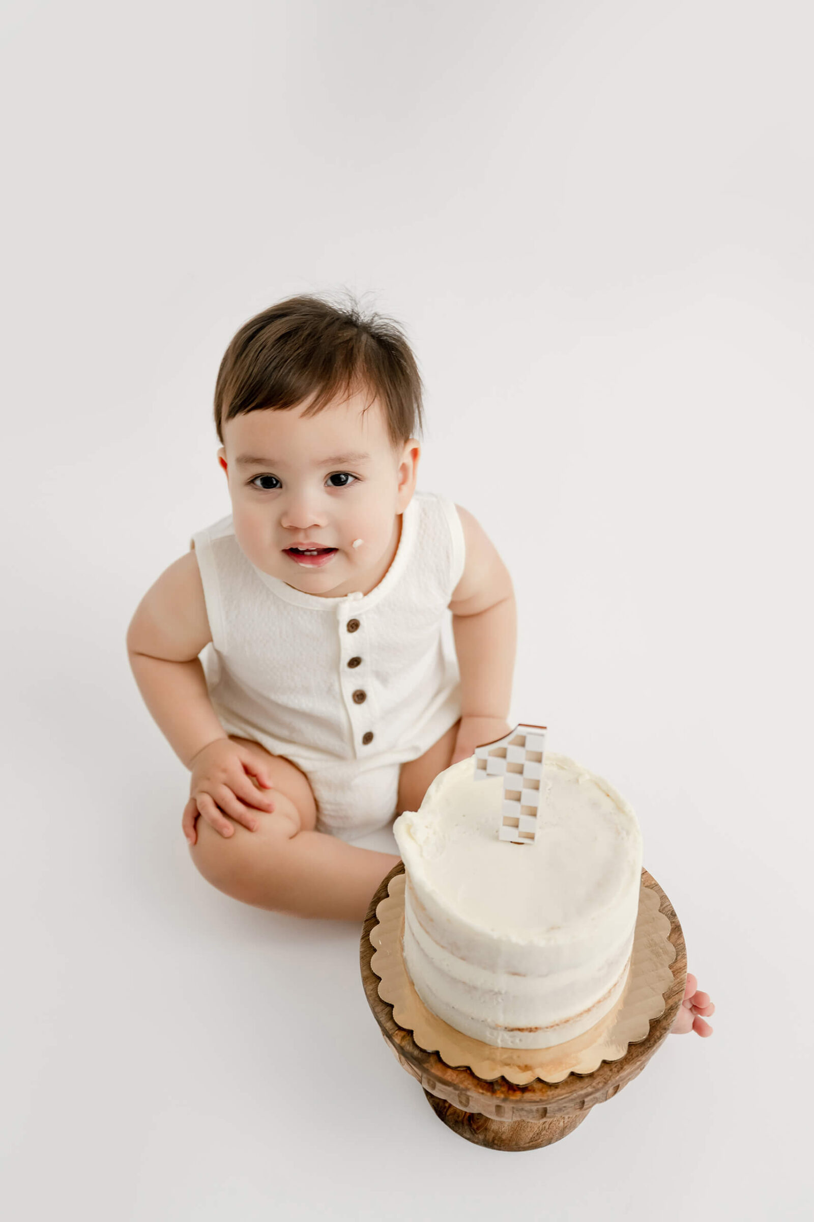 One-year-old sitting next to a white birthday cake with a little frosting on his cheek.