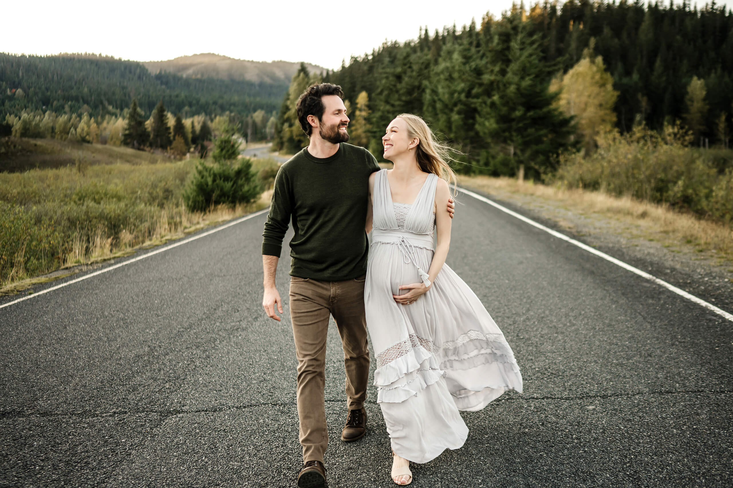 Expecting couple walking together on a quiet road with forested hills in the background.