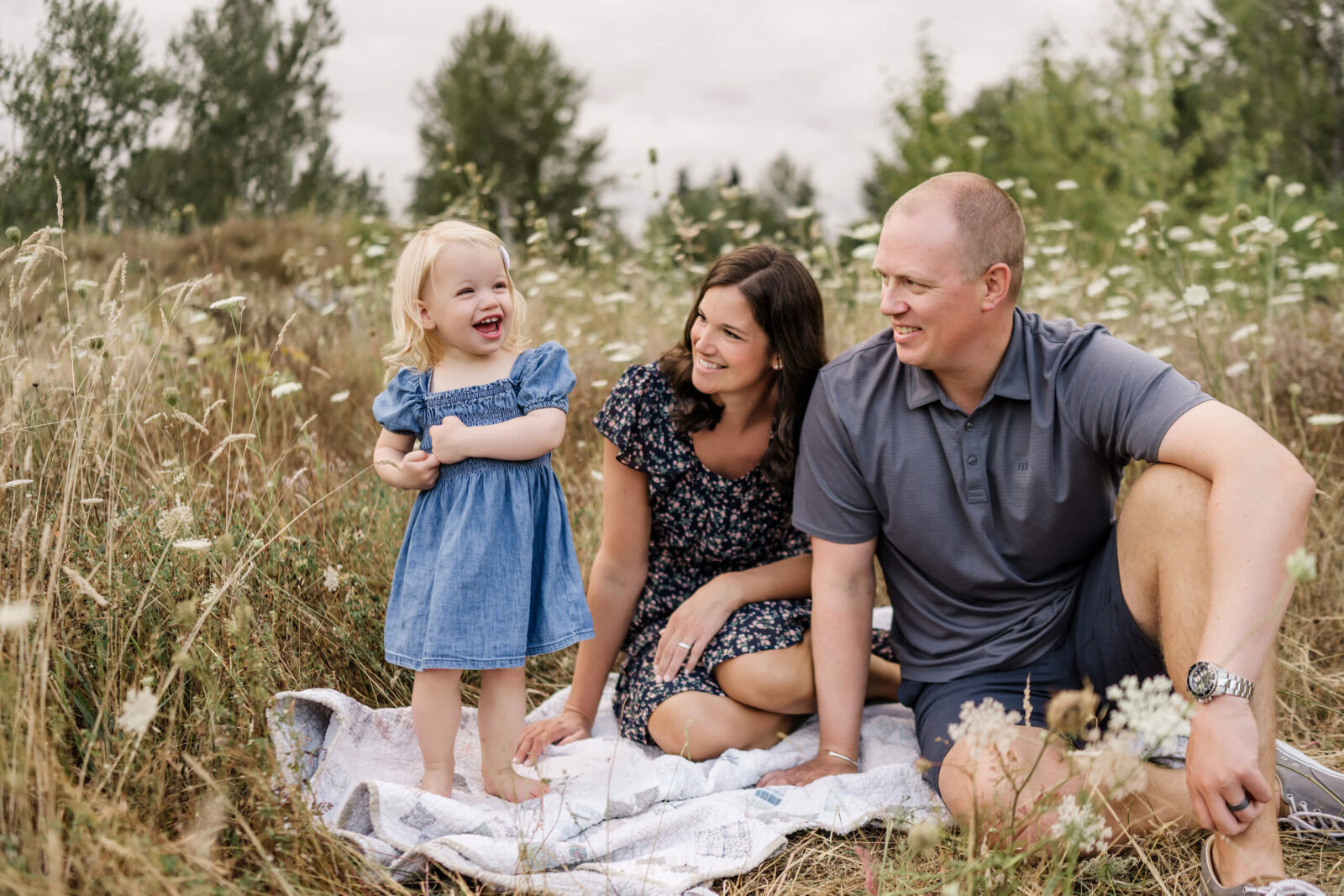 Parents sitting with their young daughter in a grassy wildflower field during an outdoor family session.
