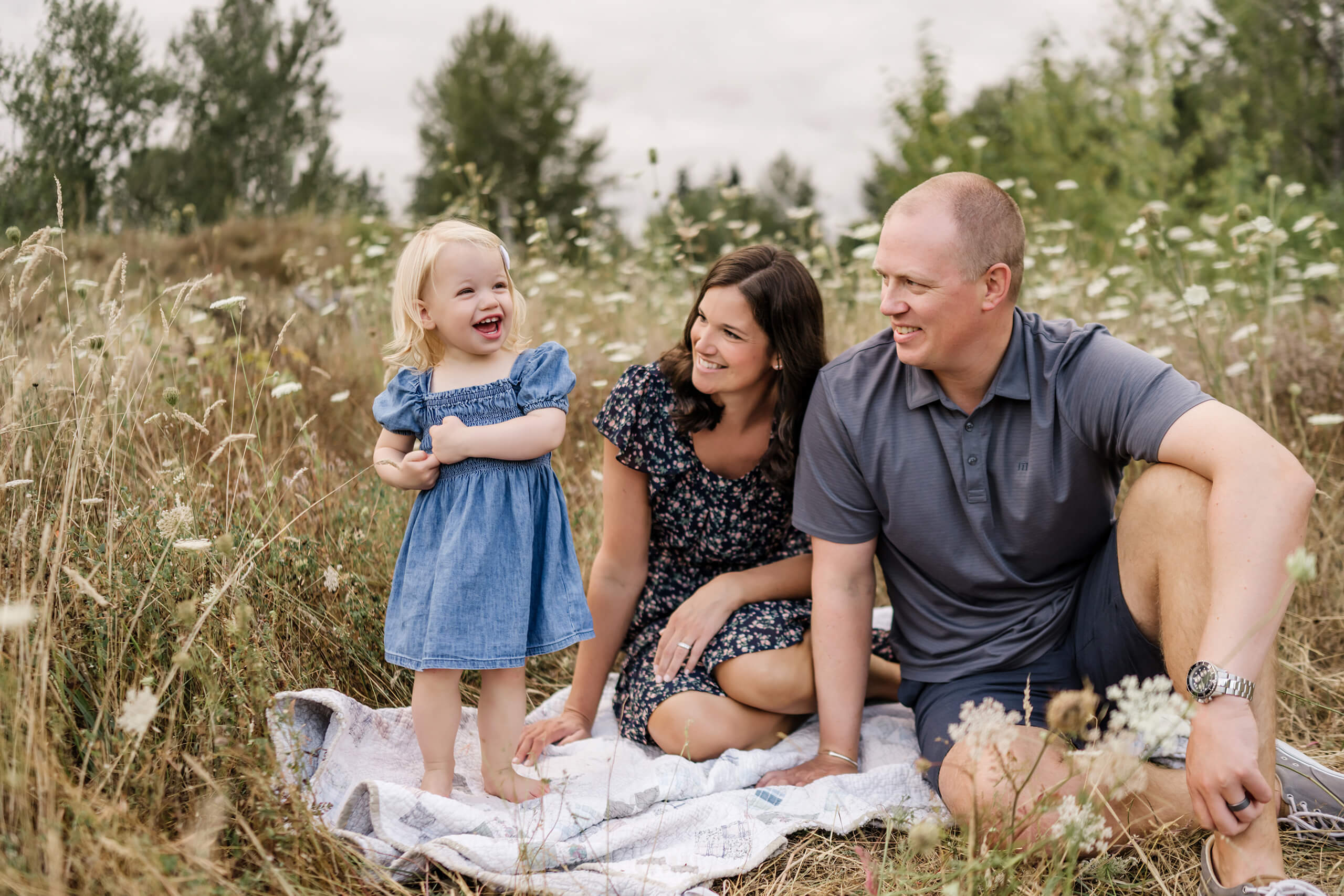 Parents sitting with their young daughter in a grassy wildflower field during an outdoor family session.