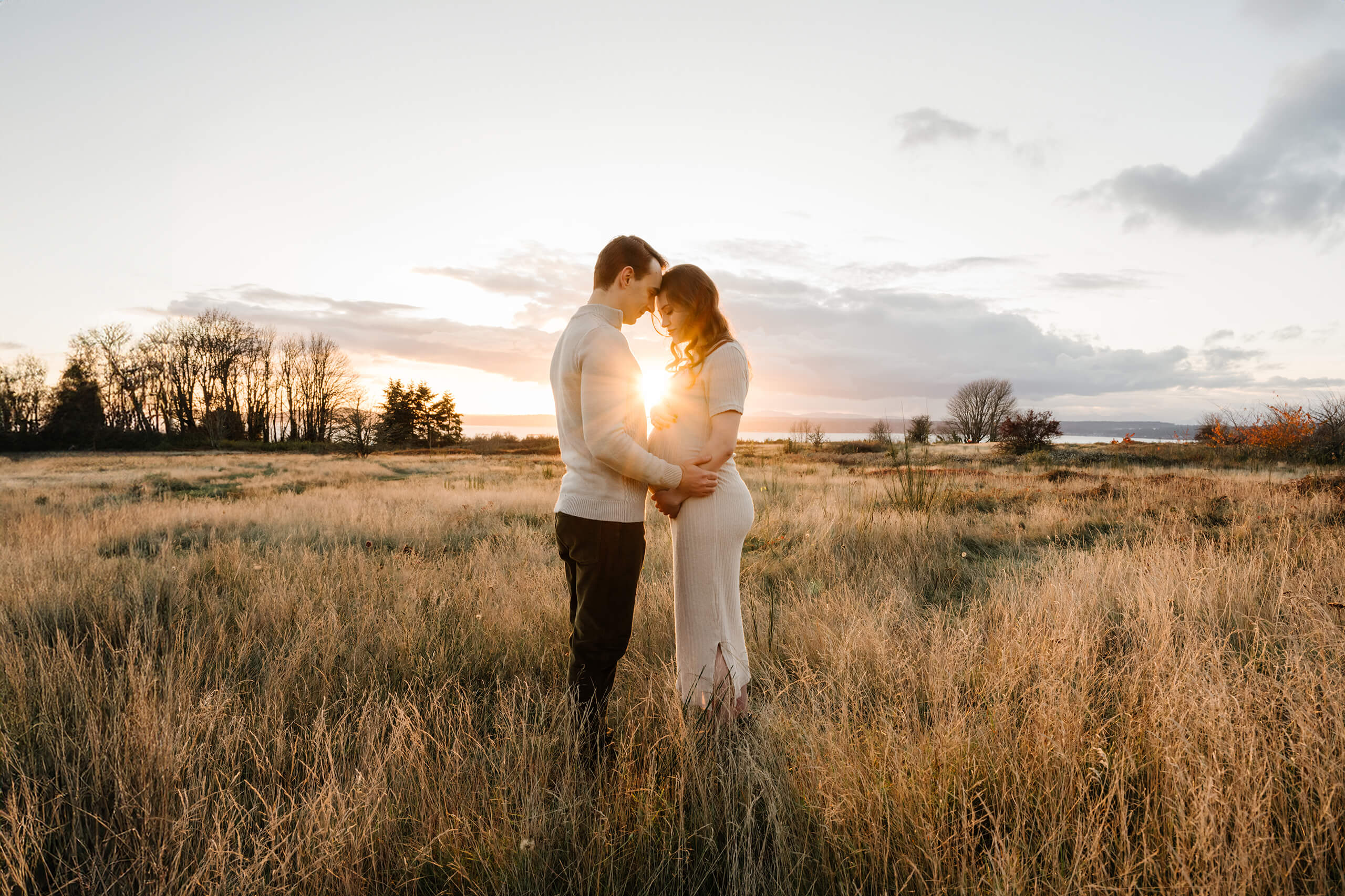 Expecting couple standing together in a golden field at sunset during a maternity photo session.