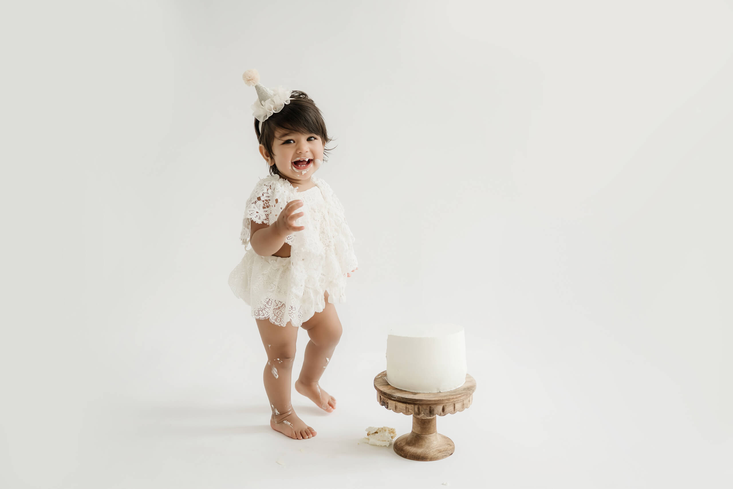 Smiling one-year-old playing with a white birthday cake during a clean studio cake smash session.