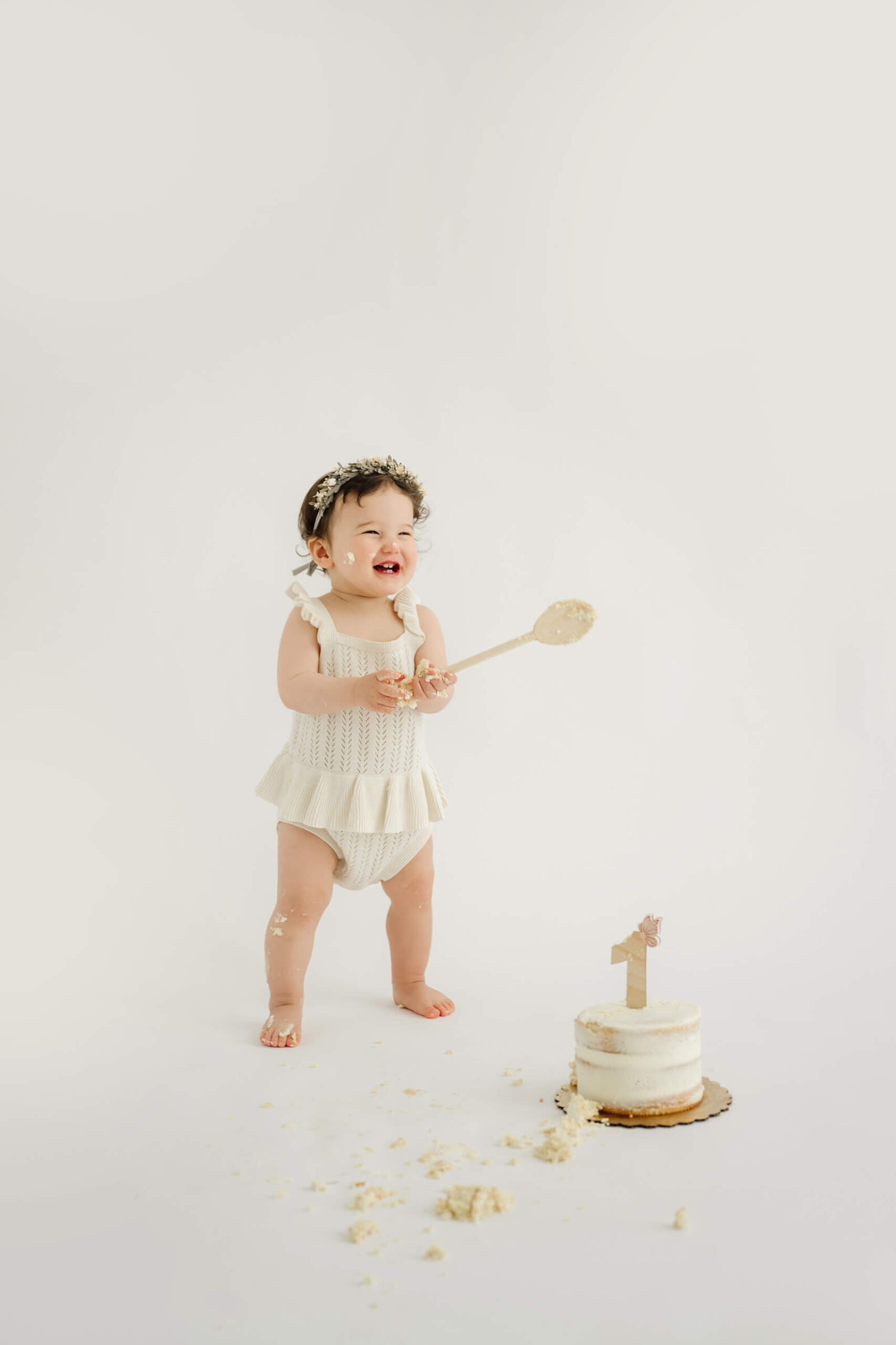 One-year-old holding a cake spoon and laughing during a playful studio cake smash session.