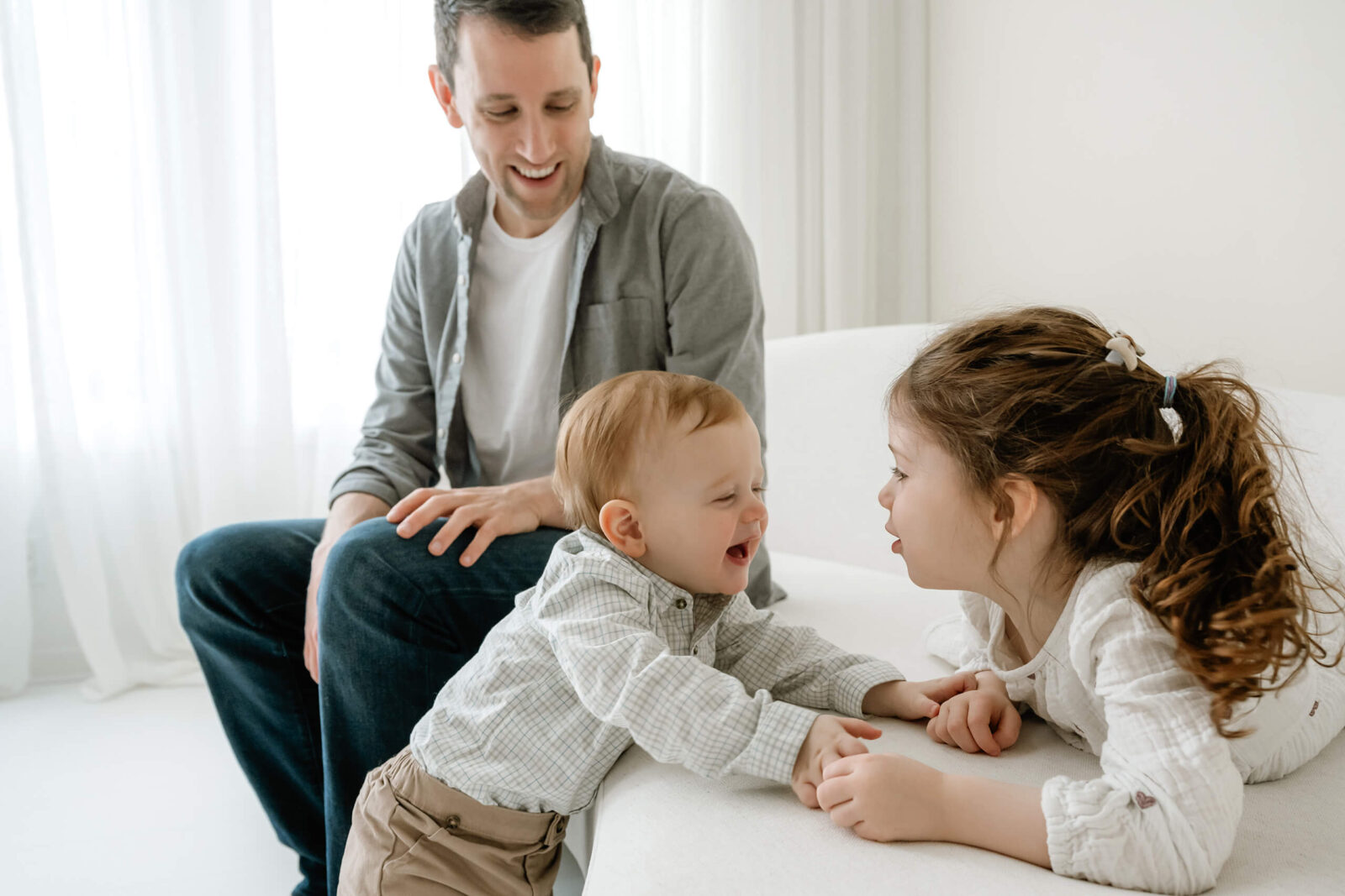 Dad watching two young children laugh together on a white couch during a studio family session.