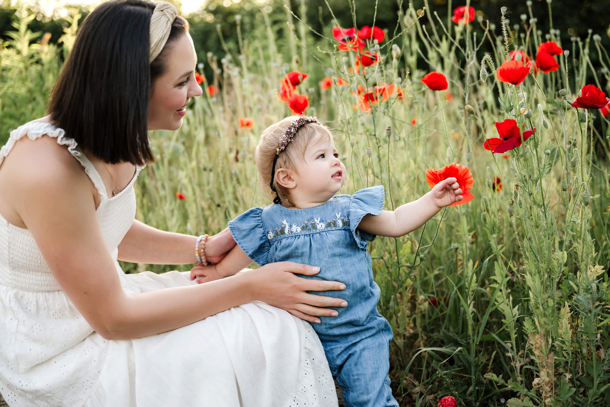 Mom sitting with her toddler daughter as she reaches toward red poppies in a flower field.
