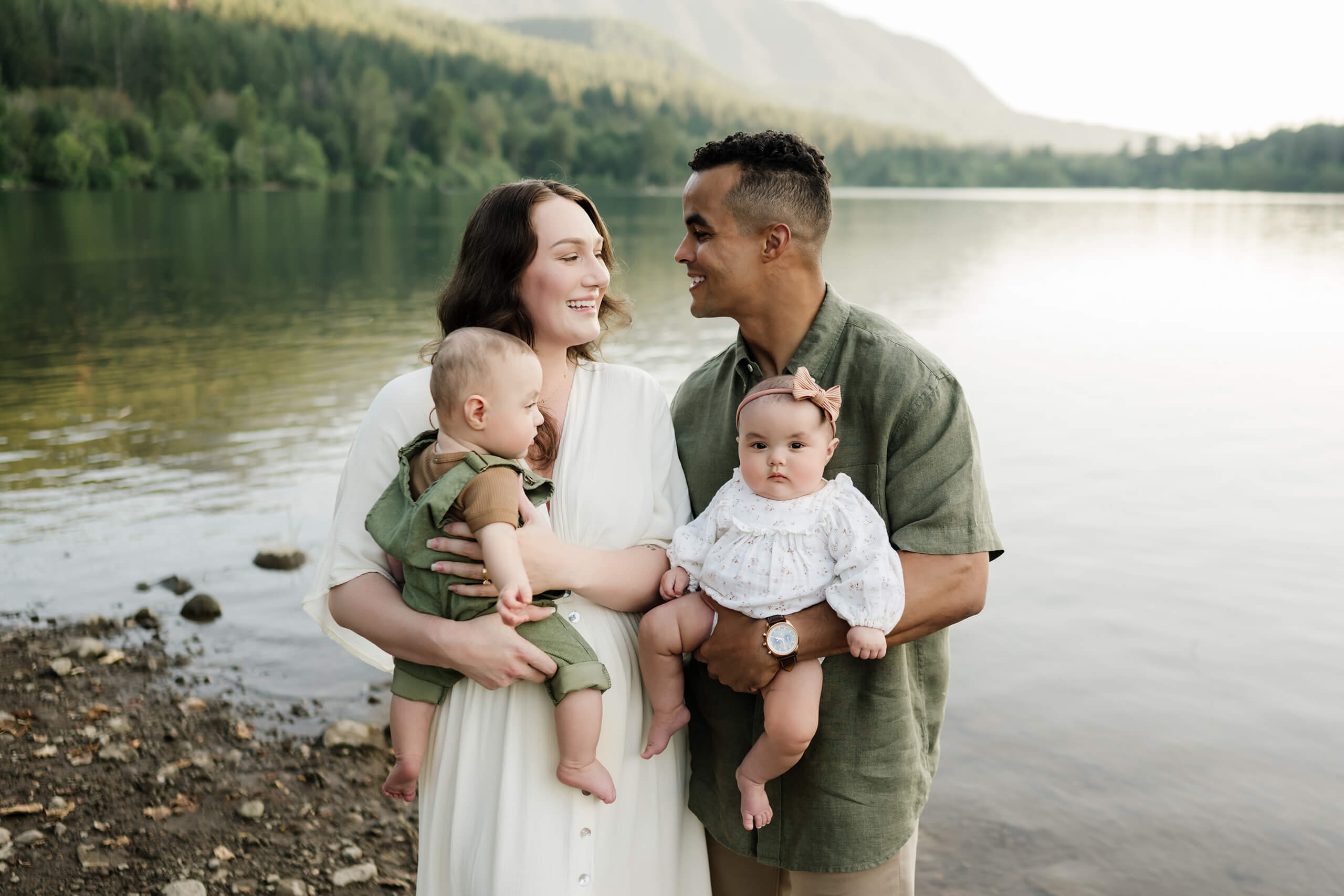Parents holding their two babies together beside a calm lake.