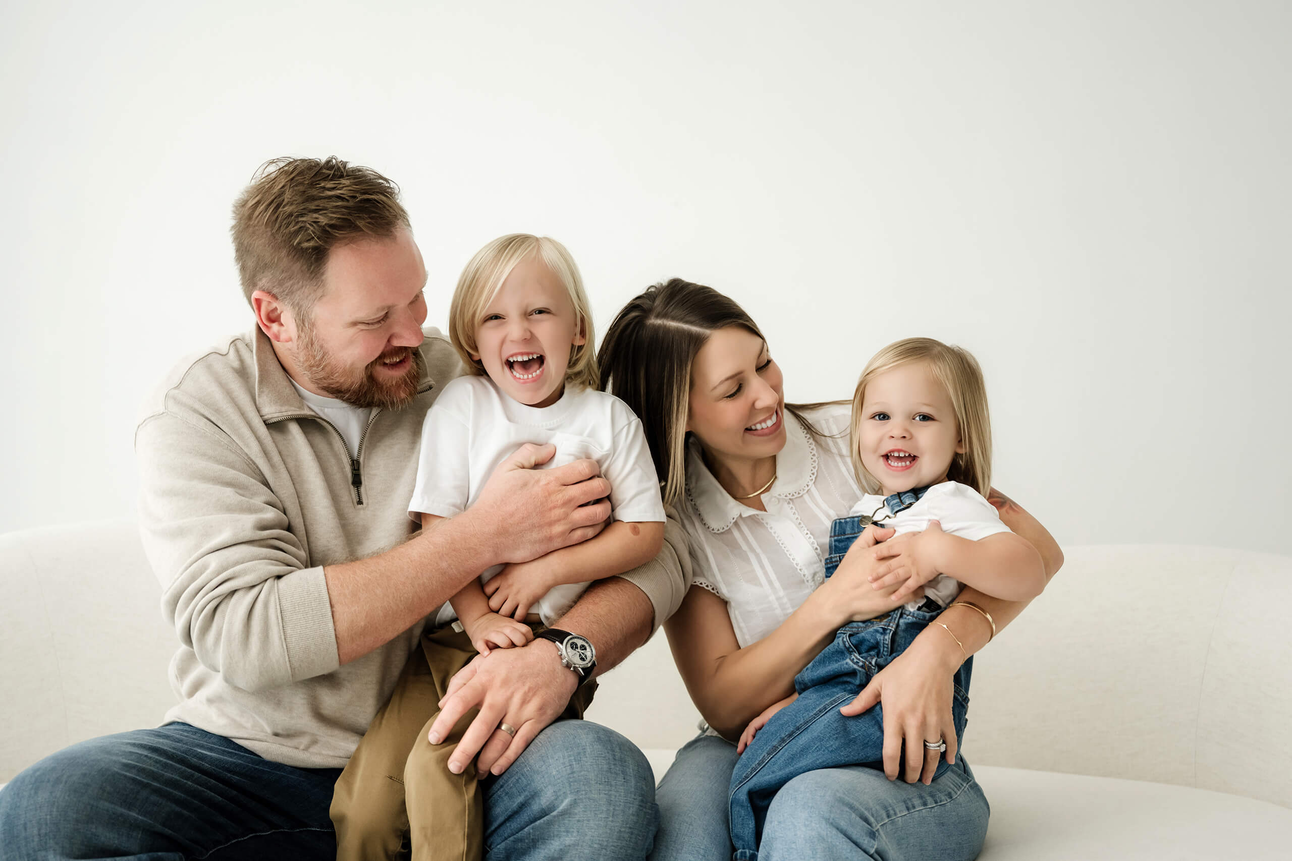 Family of four laughing together on a white couch during a studio family session.