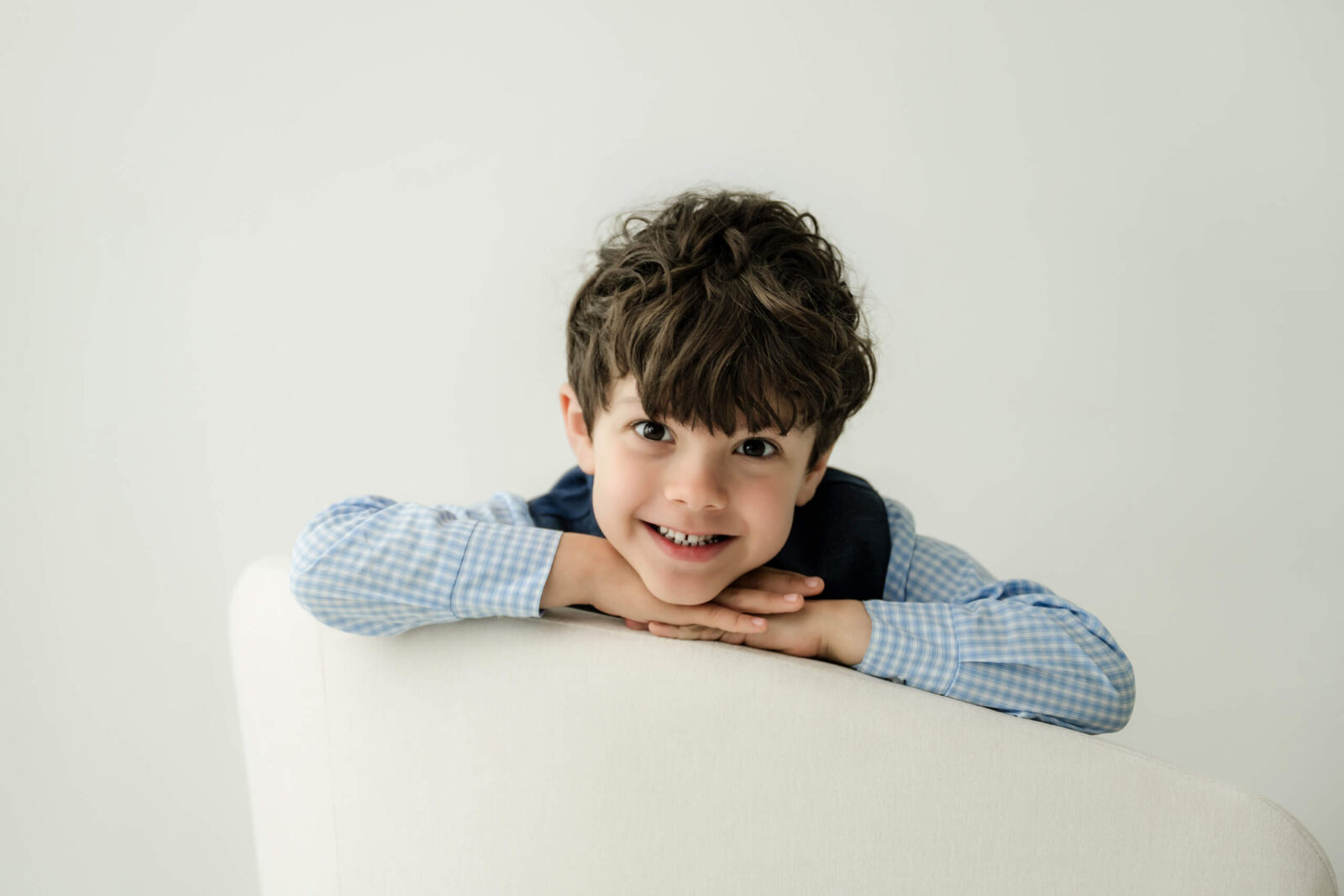 Young boy smiling over the back of a white chair during a studio family photo session.