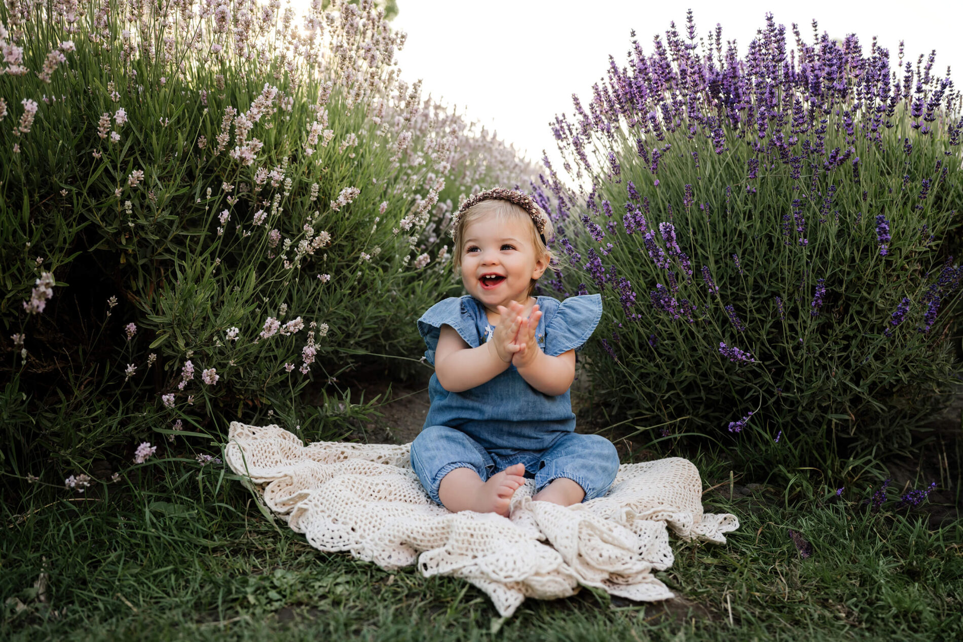 Smiling toddler sitting on a cream blanket between lavender rows during a family photo session.
