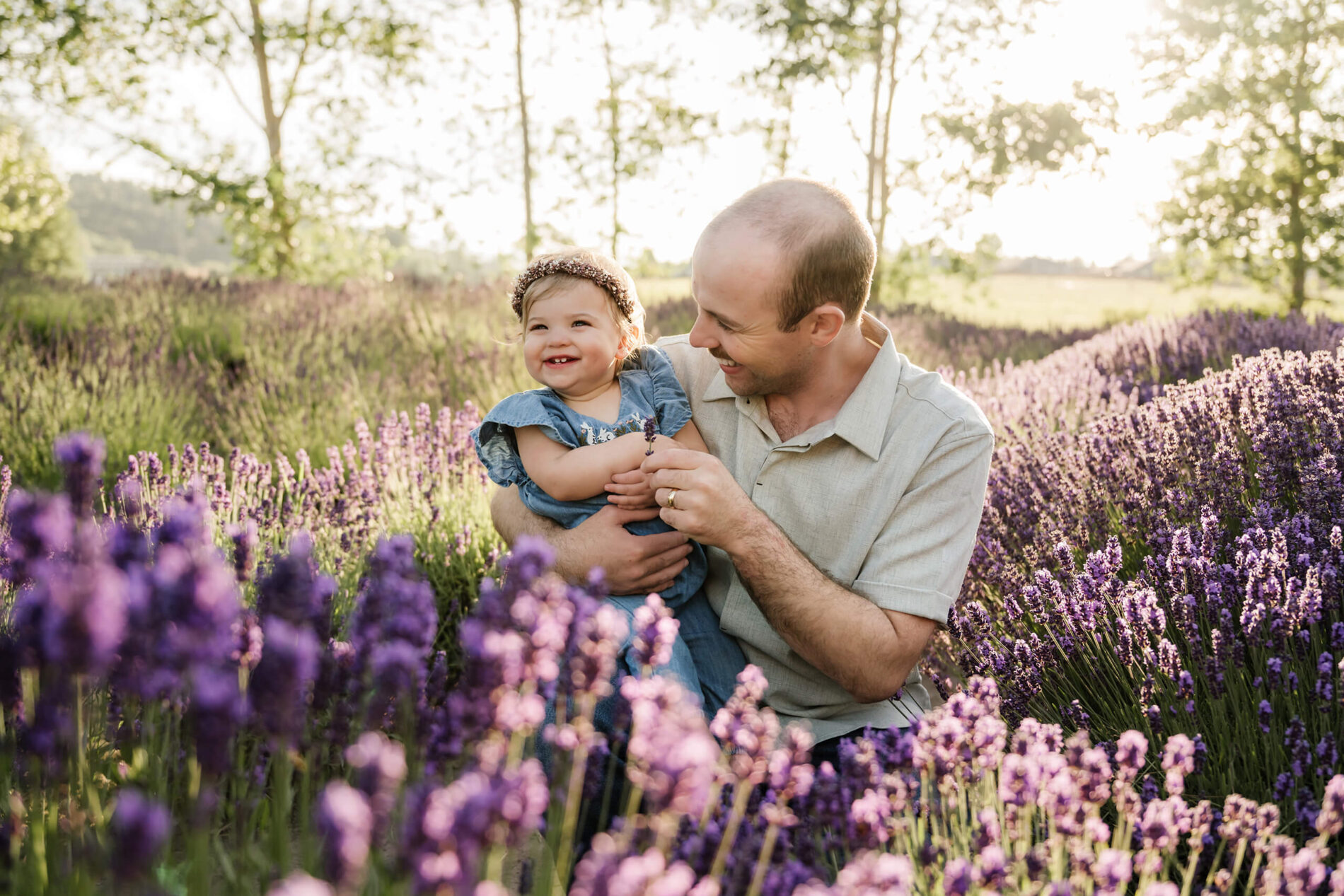 Father and toddler laughing together while sitting in purple lavender rows during golden hour.
