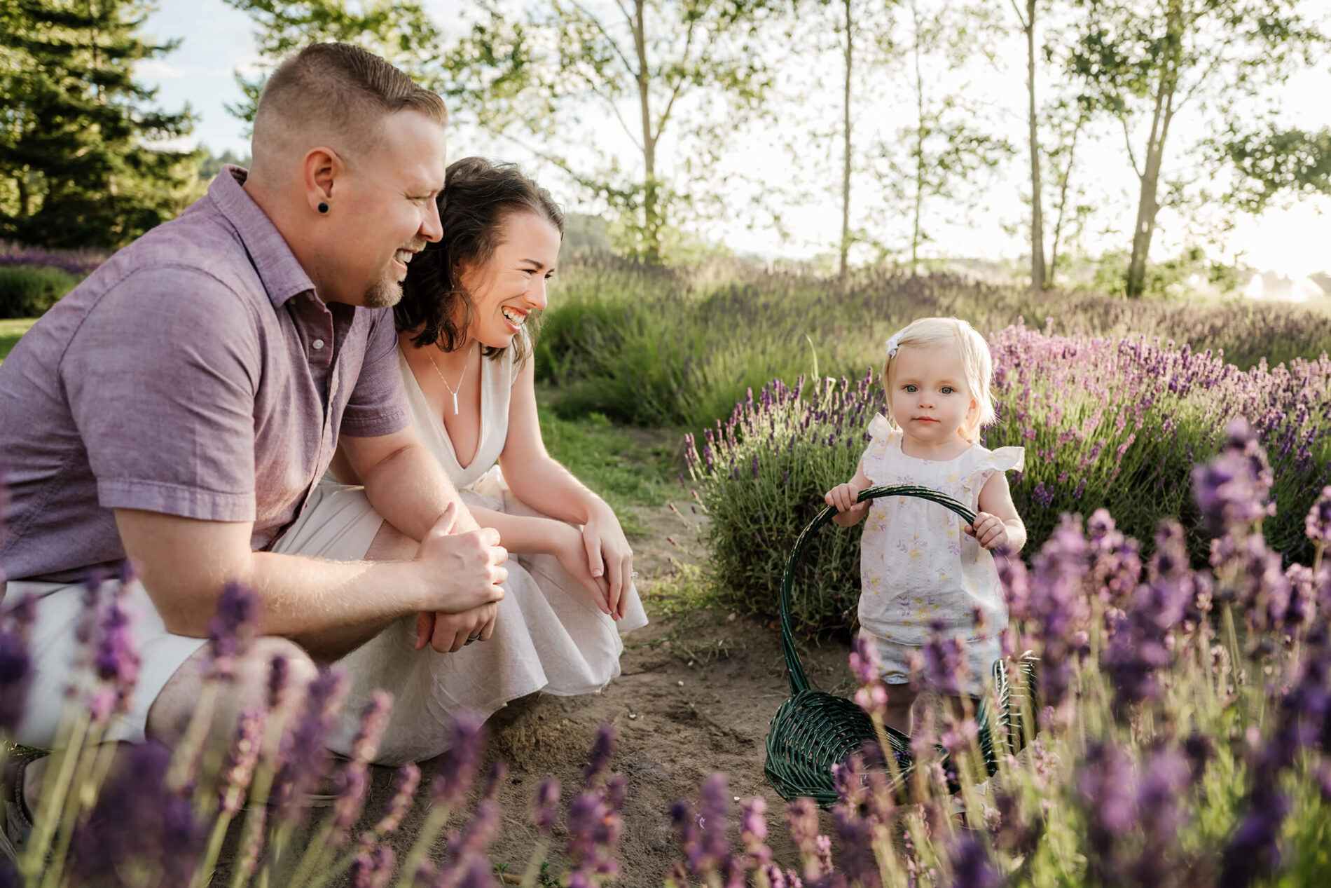 Parents smiling at their toddler as she holds a basket beside the lavender.