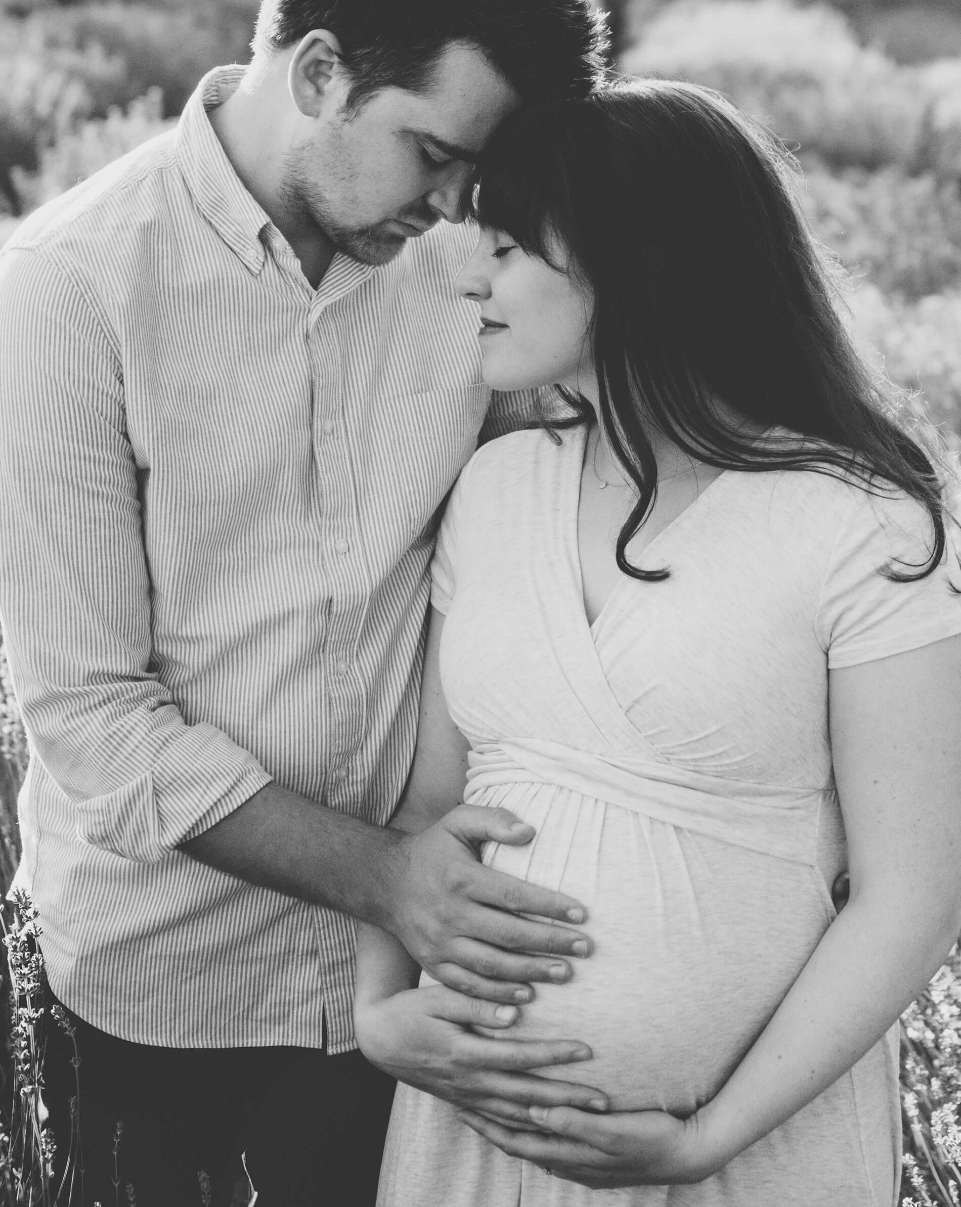 Black-and-white maternity portrait of expecting parents holding mom’s belly in a lavender field.