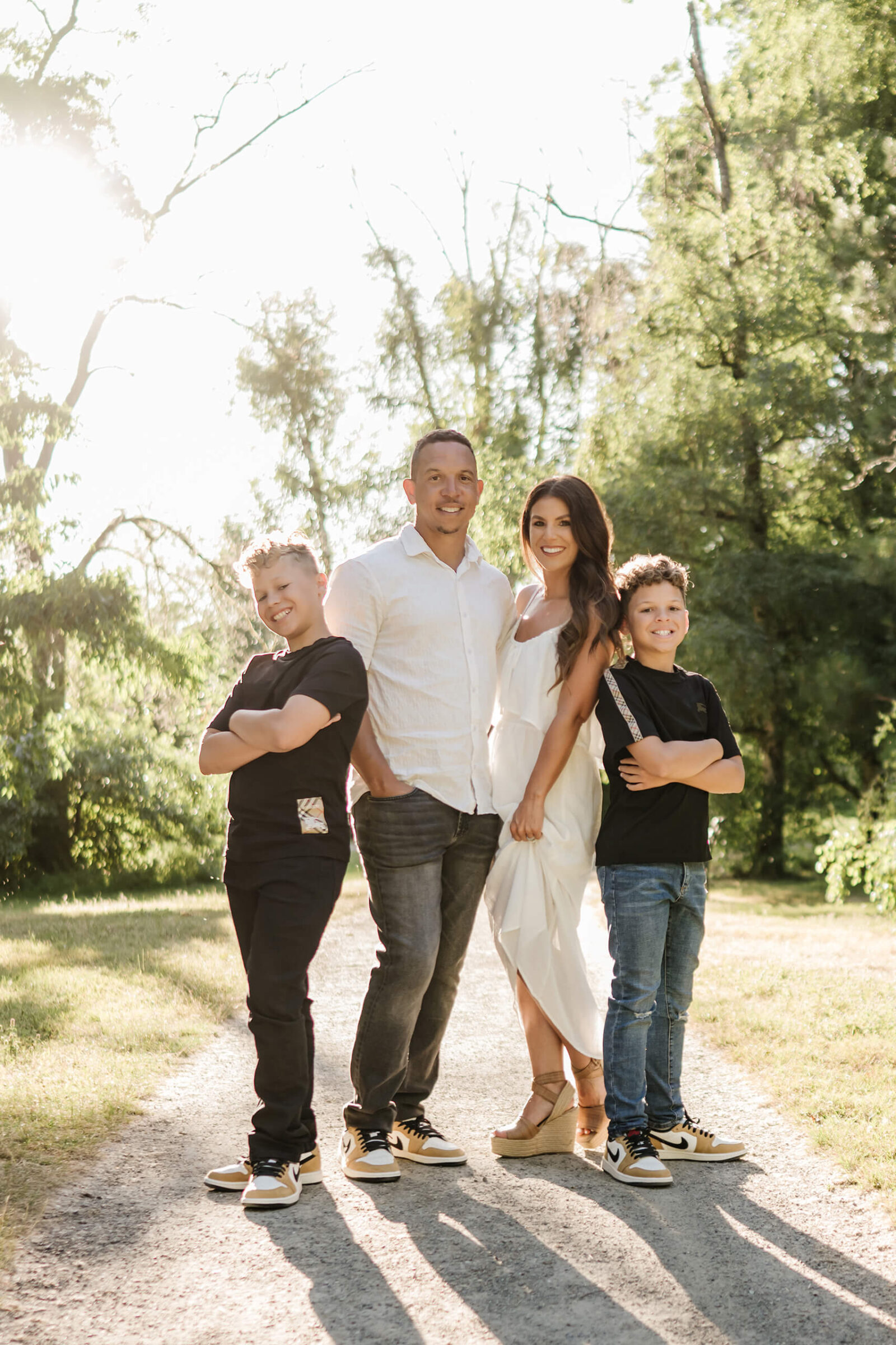 Family of four standing together on a sunlit tree-lined path during an outdoor family session.