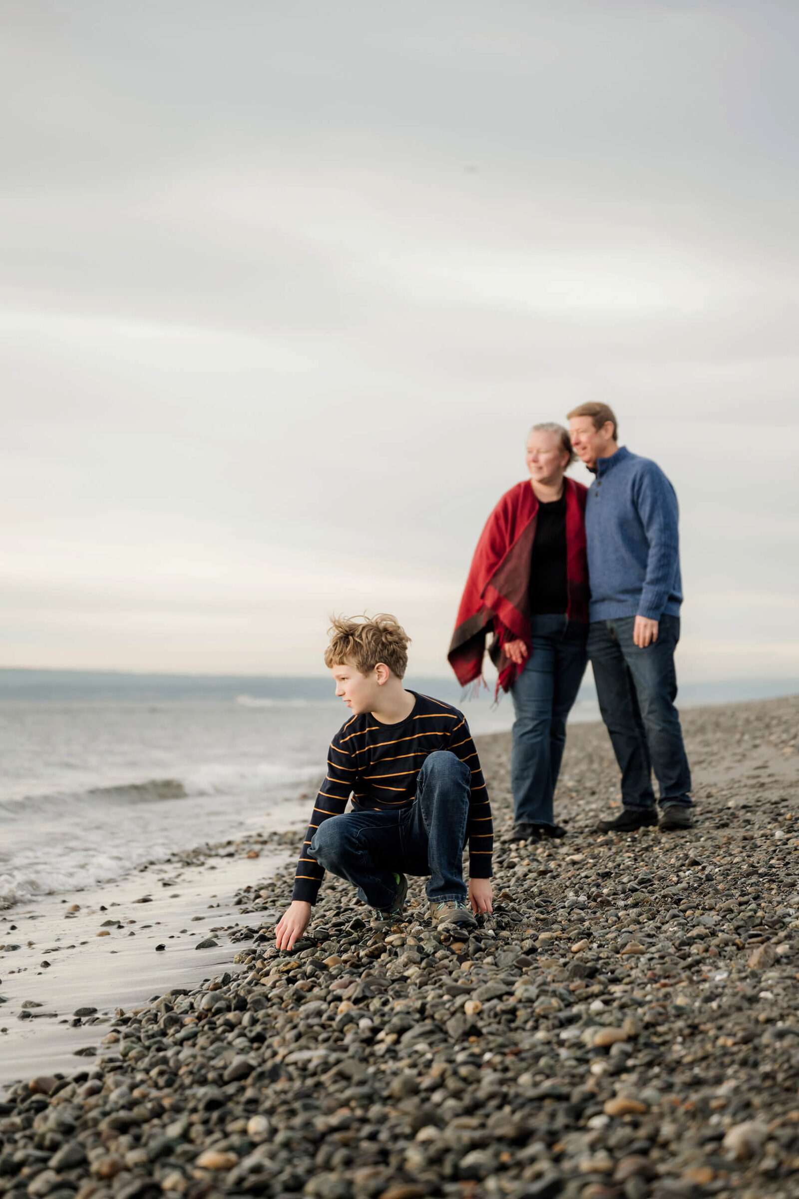 Boy crouching on a rocky beach while his parents stand together in the background.