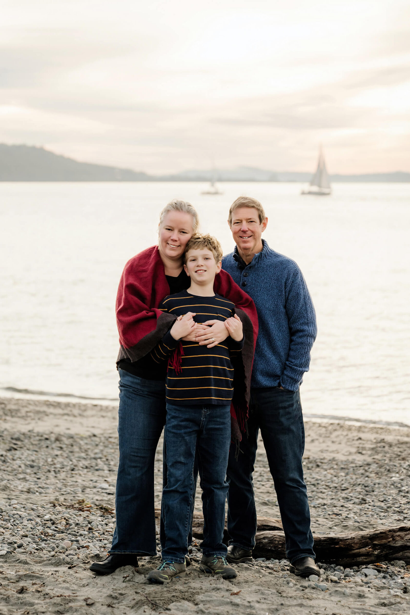 Parents and son standing together on a rocky beach with sailboats in the background.