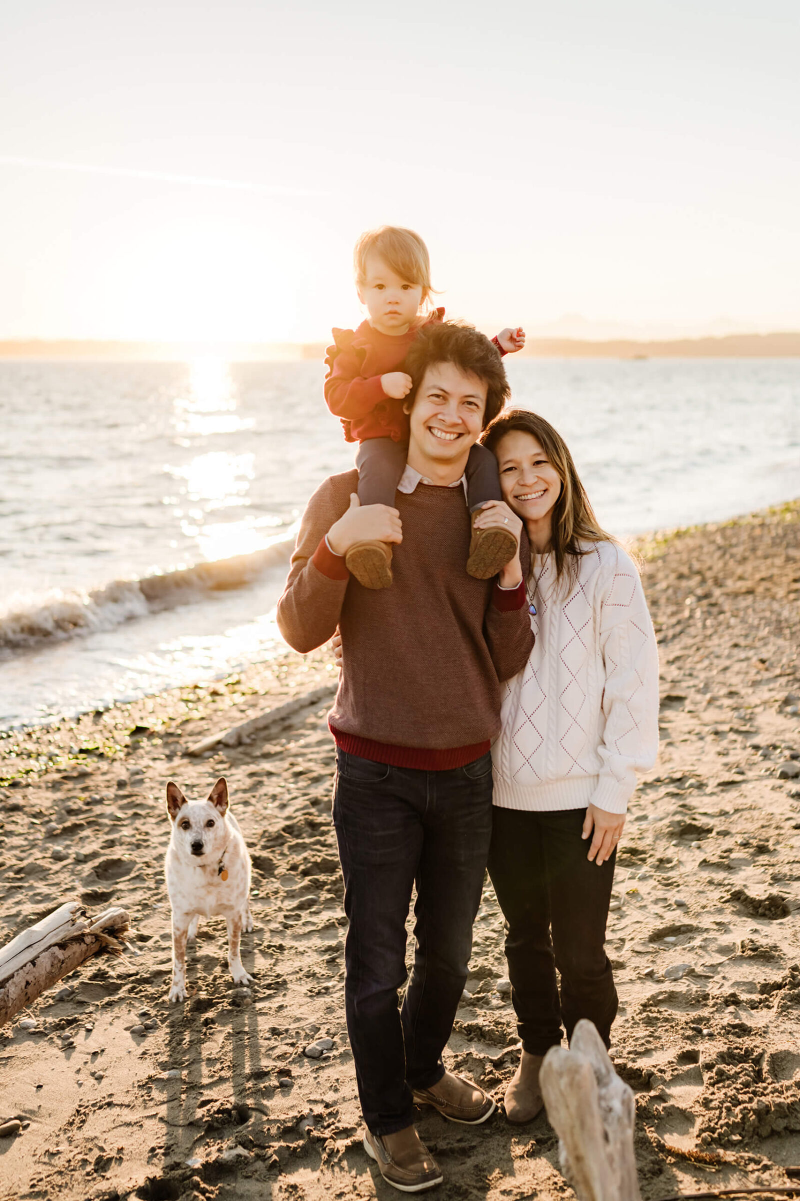 Family of three standing with their dog on a sandy beach in golden evening light.